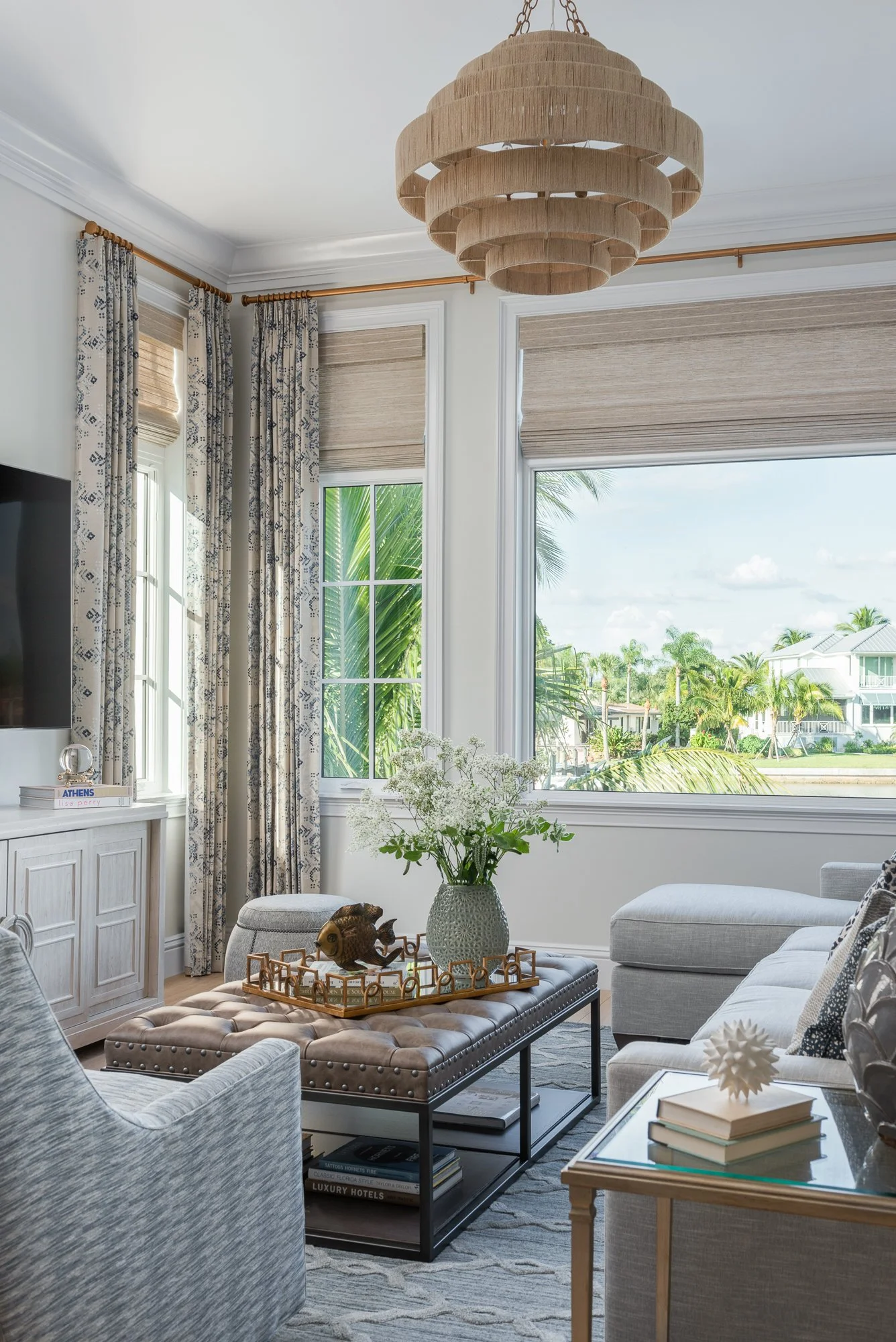 Living room with large windows, beige curtains, and a chandelier. Contains a beige tufted ottoman with decorative items, a side table with books and a white sculpture, and light-colored furniture.