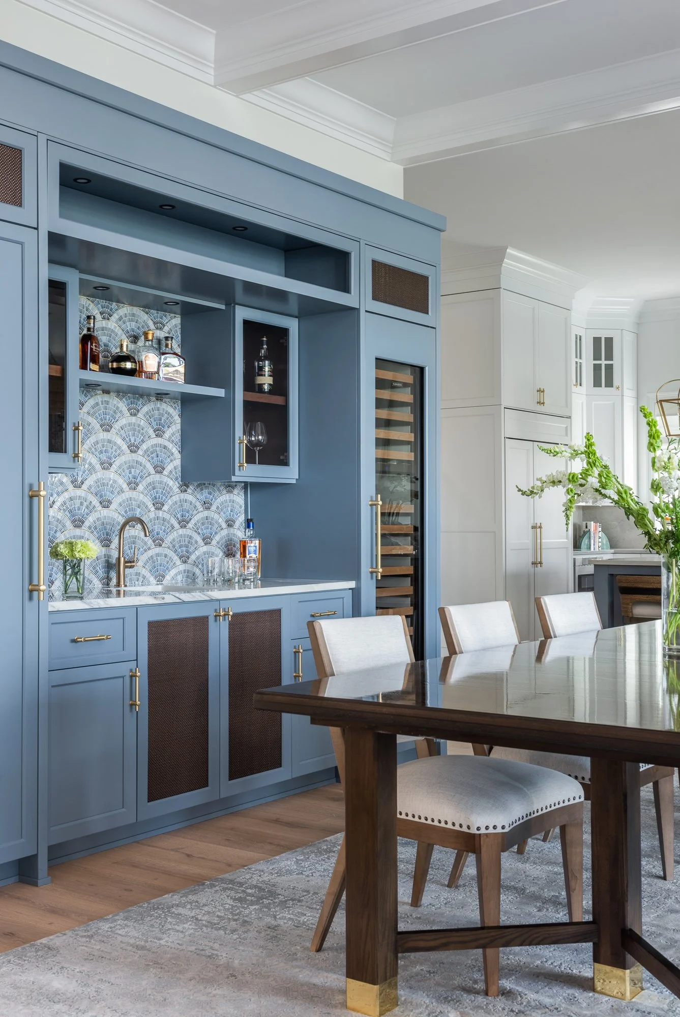 A kitchen with a blue cabinet area featuring a wine fridge, bar supplies, and bottles of alcohol, with a marble countertop, a sink with a faucet, and open shelving. There is a wooden dining table with beige upholstered chairs, a large glass vase with greenery, and a white background with built-in white cabinets.