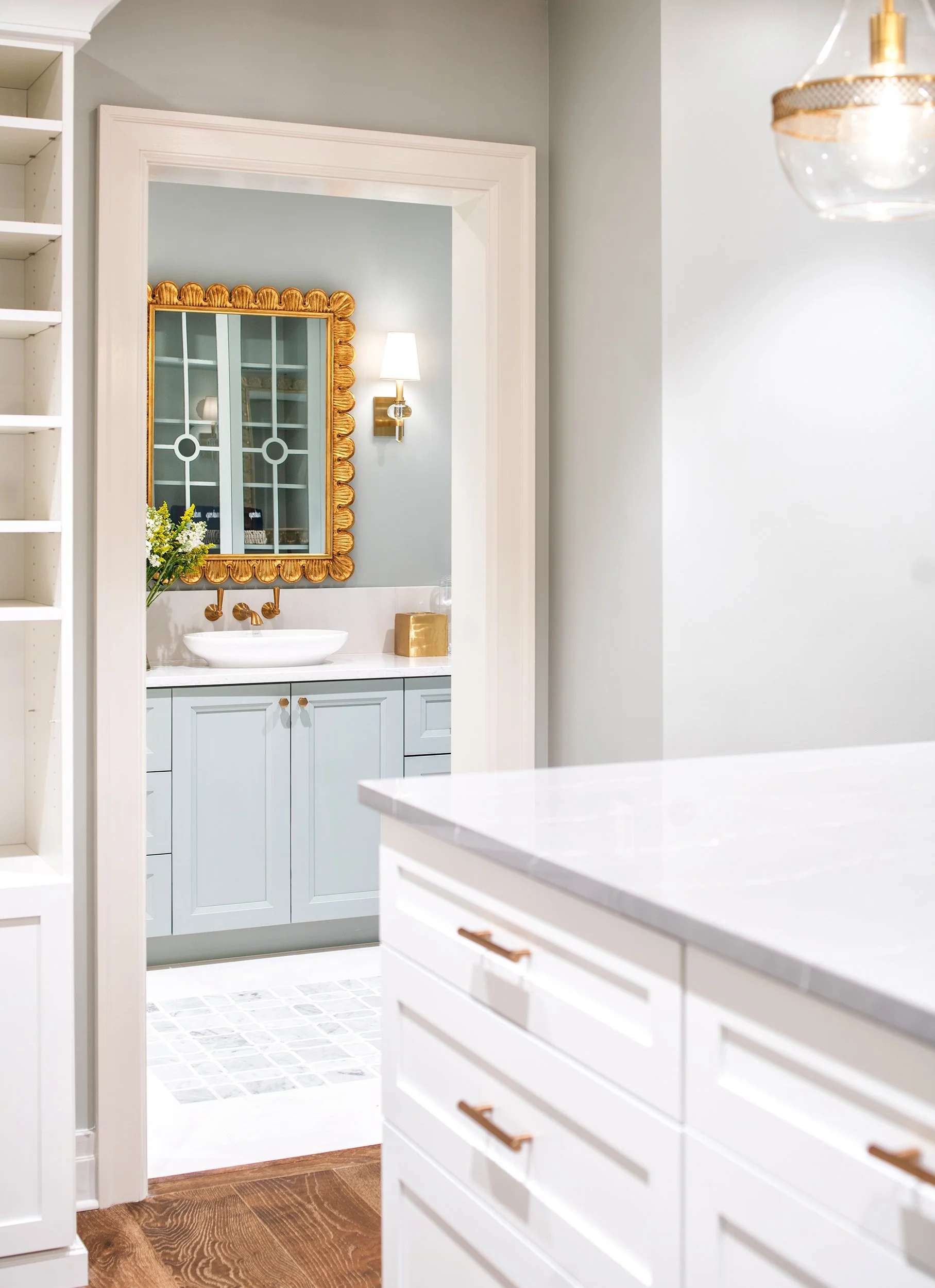 View of a bathroom seen through a doorway, featuring a light blue vanity, a white vessel sink, gold fixtures, a gold-framed mirror, and a wall-mounted light fixture, with a small bouquet of flowers on the vanity.