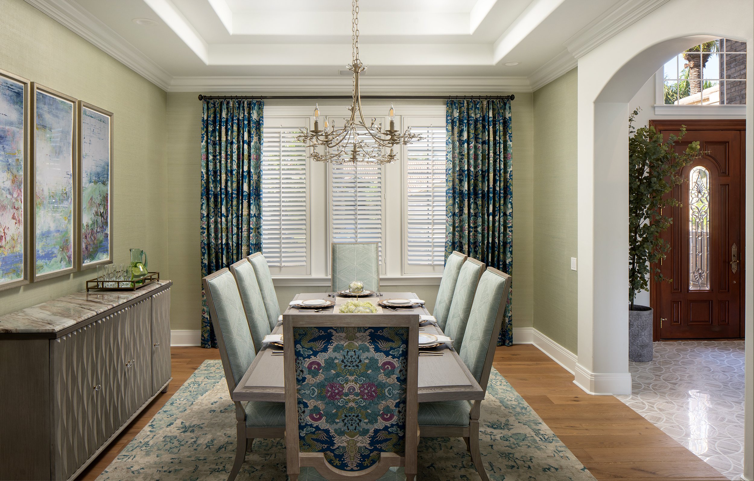 Dining room with a wooden dining table and eight upholstered chairs, a chandelier overhead, large windows with shutters and floral curtains, artwork on the wall, a sideboard with decorative items, and an entryway with a wooden door and indoor plant.
