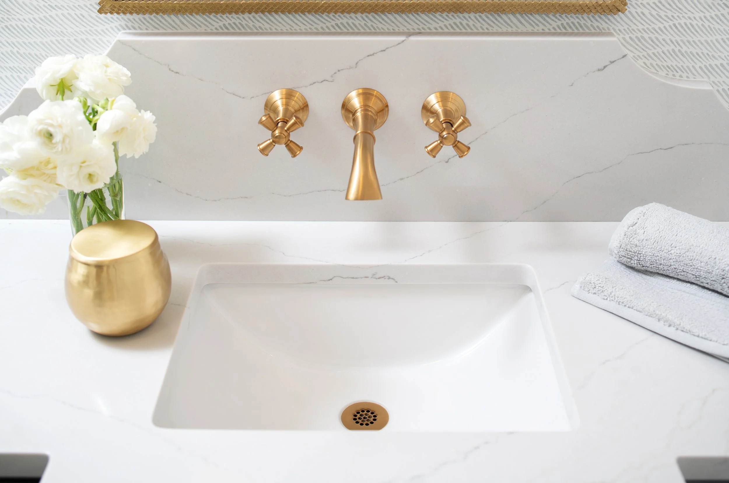 Bathroom sink with gold faucet and handles, white marble countertop, a vase with white flowers, two folded gray towels.
