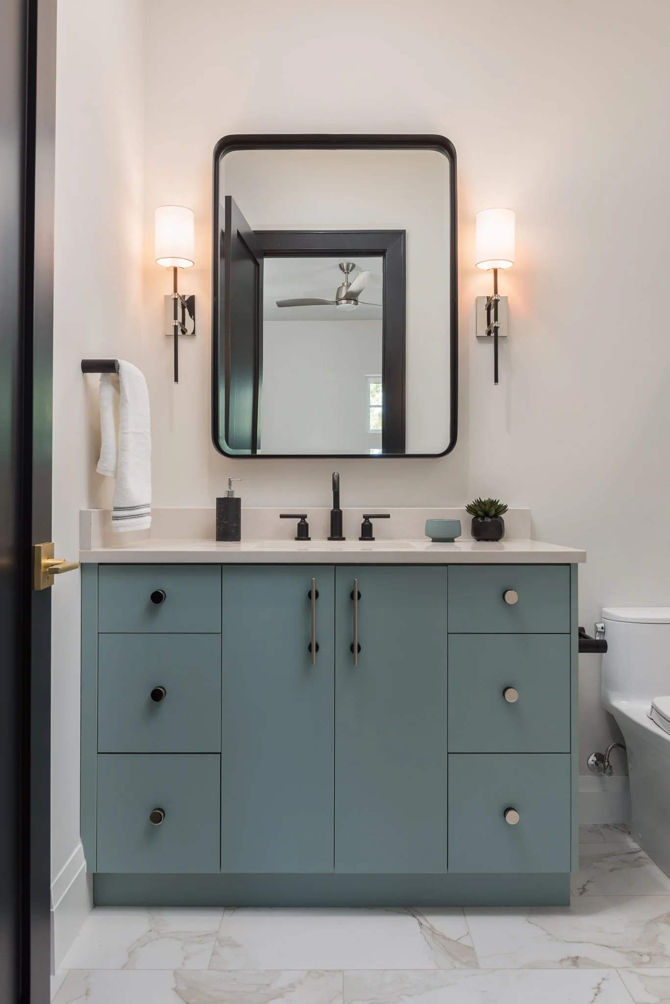 Modern bathroom vanity with a black-framed mirror, light fixtures, and a small potted plant on the countertop.
