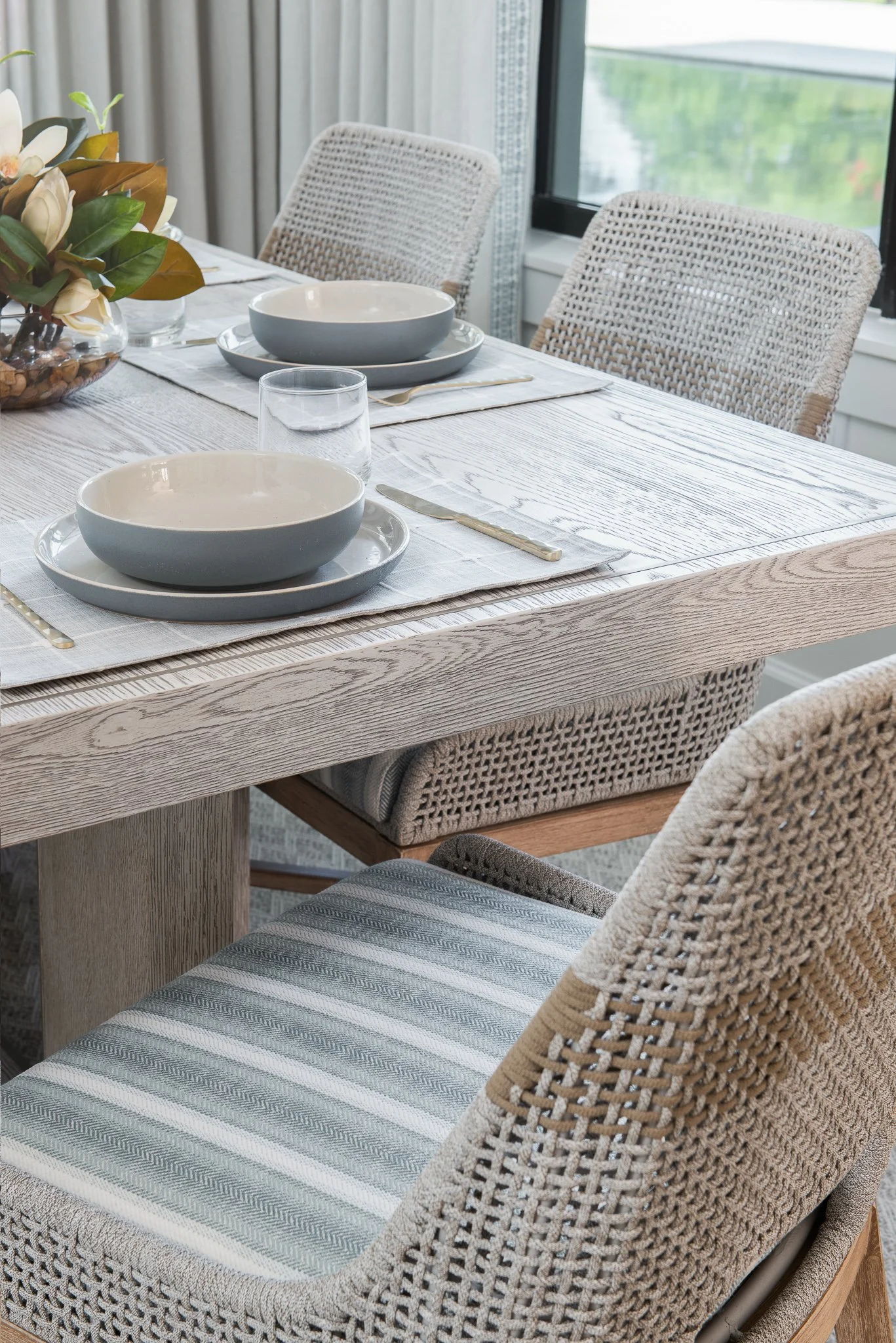 A dining table set for a meal with bowls, plates, a glass of water, and cutlery, with woven chairs and a vase of flowers nearby.