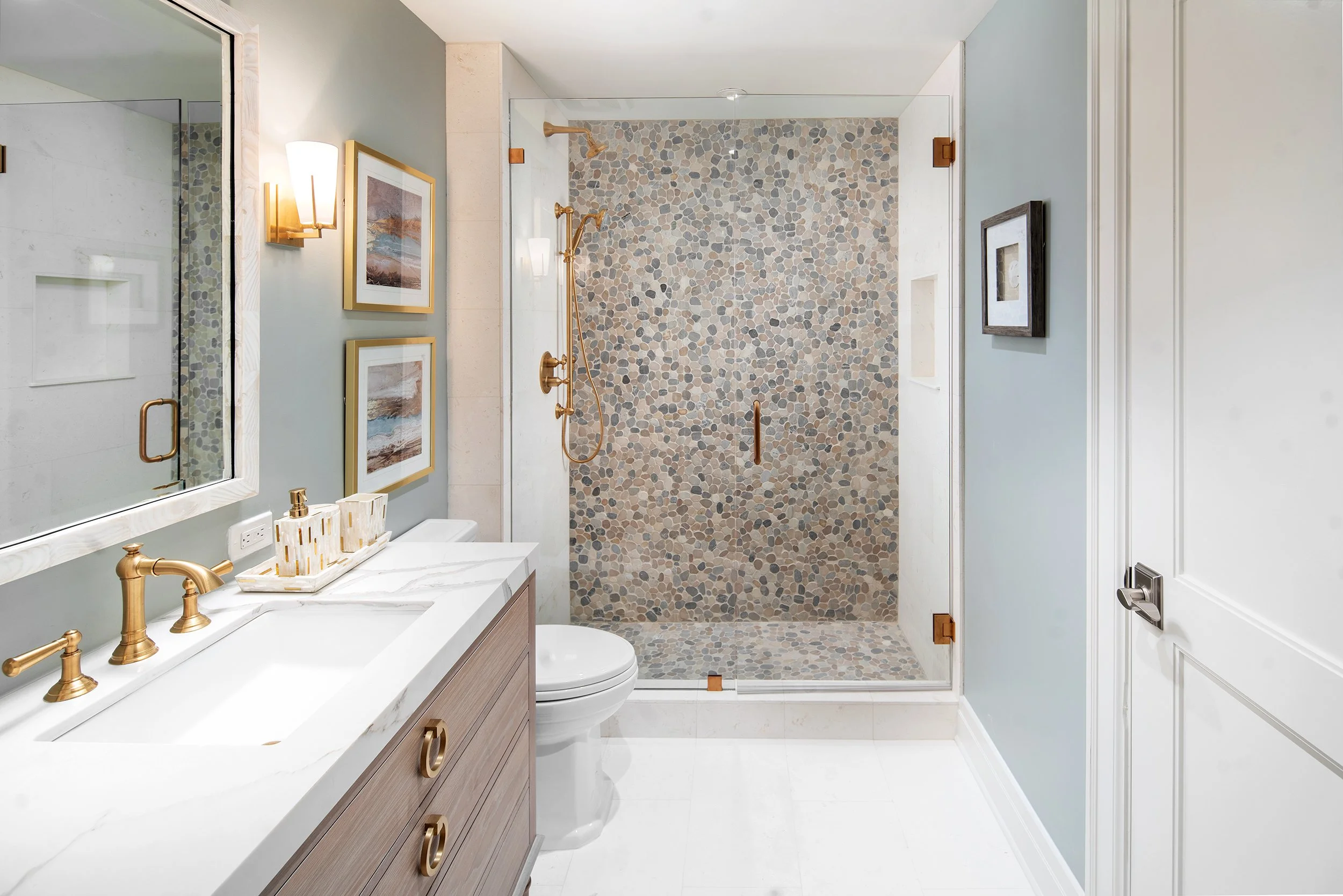 A modern bathroom featuring a glass-enclosed pebble-textured shower, light blue walls, gold fixtures, a wooden vanity with a marble countertop, framed artwork on the walls, and a white door.