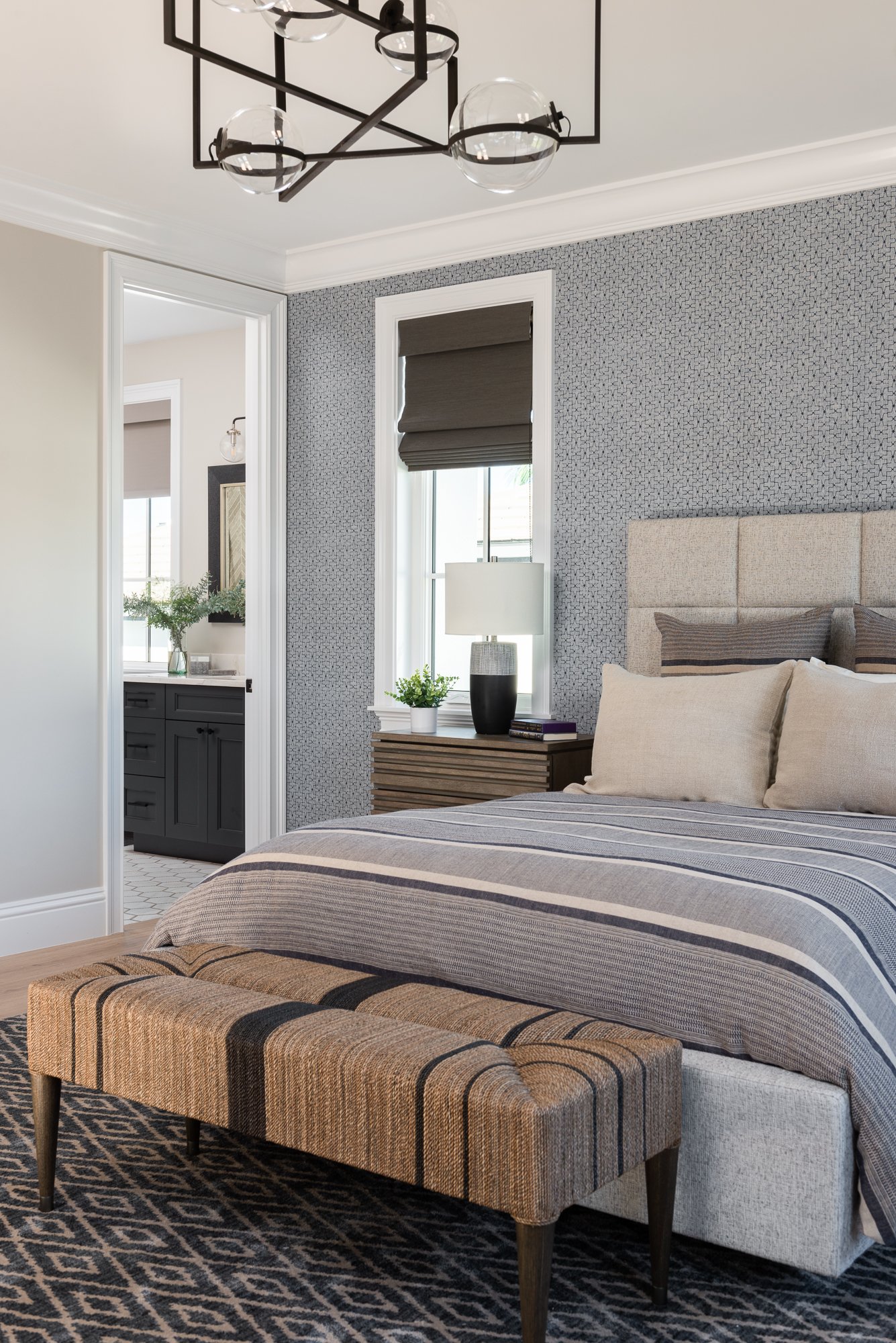 A bedroom with a bed, striped bedding, a padded headboard, a striped upholstered bench at the foot, a nightstand with a modern lamp, a small plant, and books. There is a nearby bathroom with dark cabinetry and a mirror.