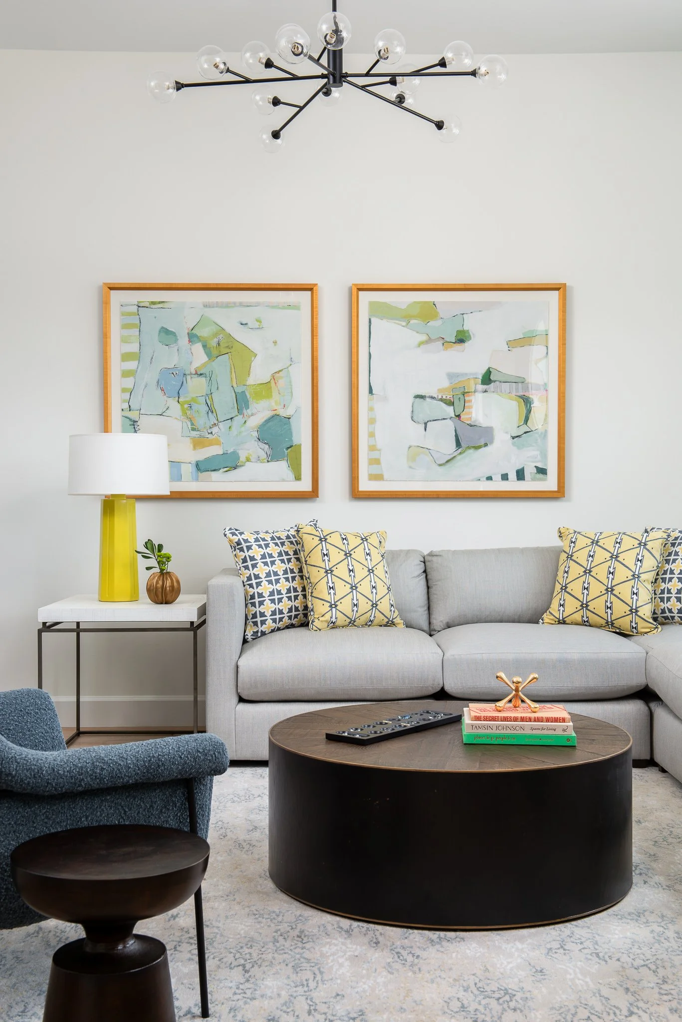 Living room with a gray sectional sofa, yellow and navy patterned pillows, a black oval coffee table with books and a decorative piece, a white side table with a yellow lamp and small plant, abstract landscape paintings on the wall, a white textured 