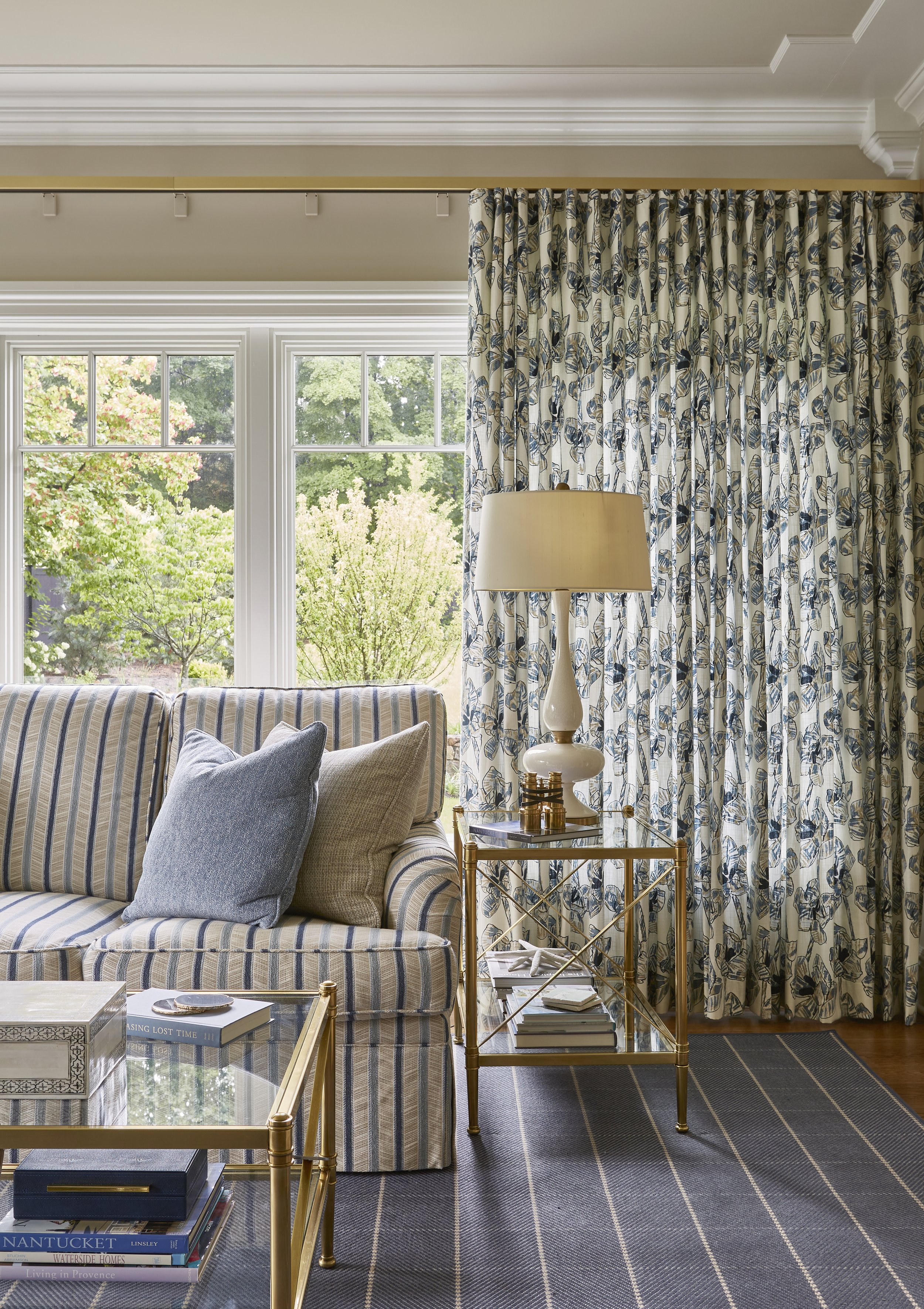 Living room with striped sofa, blue and beige pillows, gold side table with books, and patterned curtains by large windows showing green trees outside.