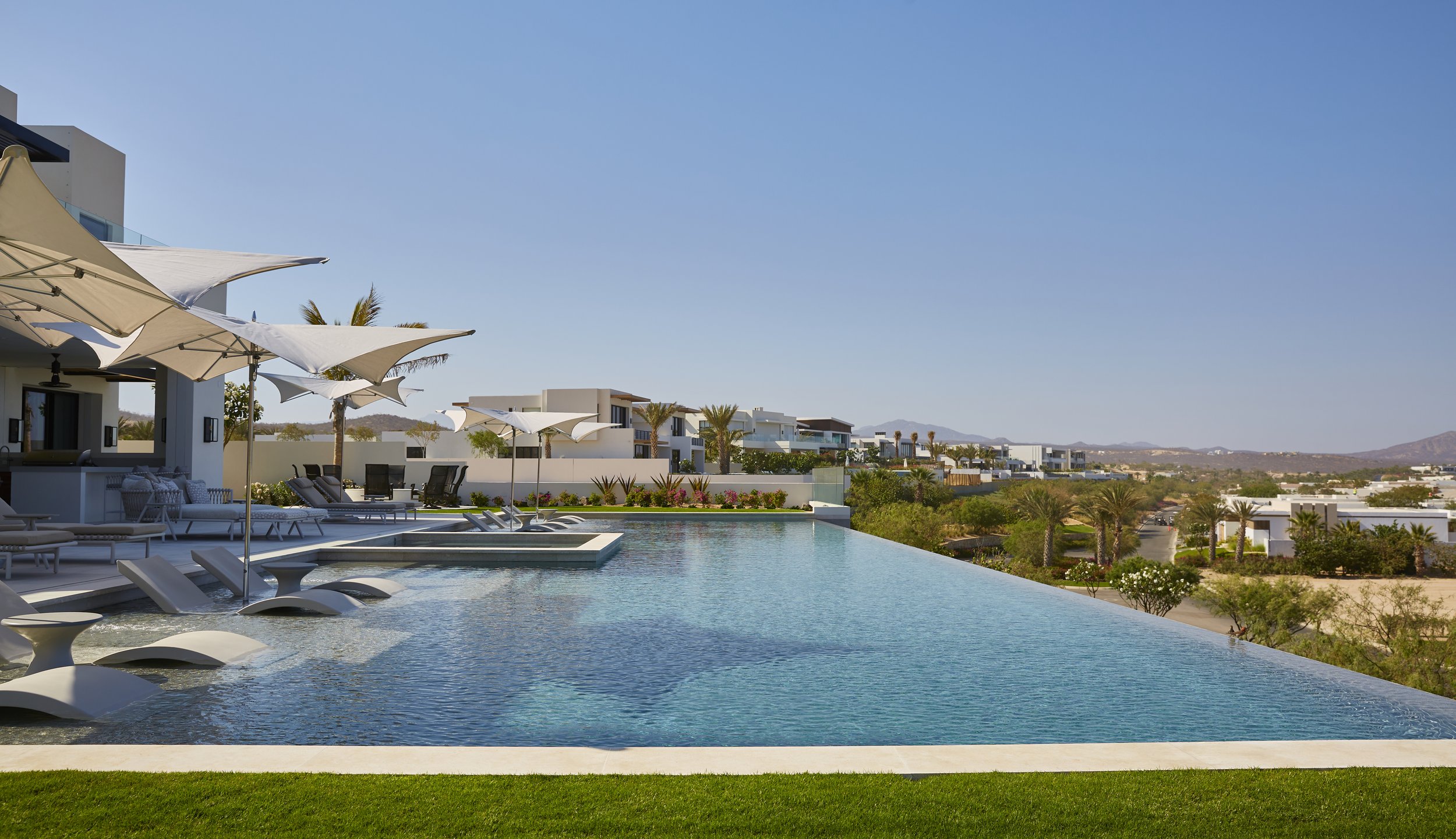 Luxury outdoor pool with sun loungers, umbrellas, modern houses, palm trees, and a desert landscape in the distance under a clear blue sky.