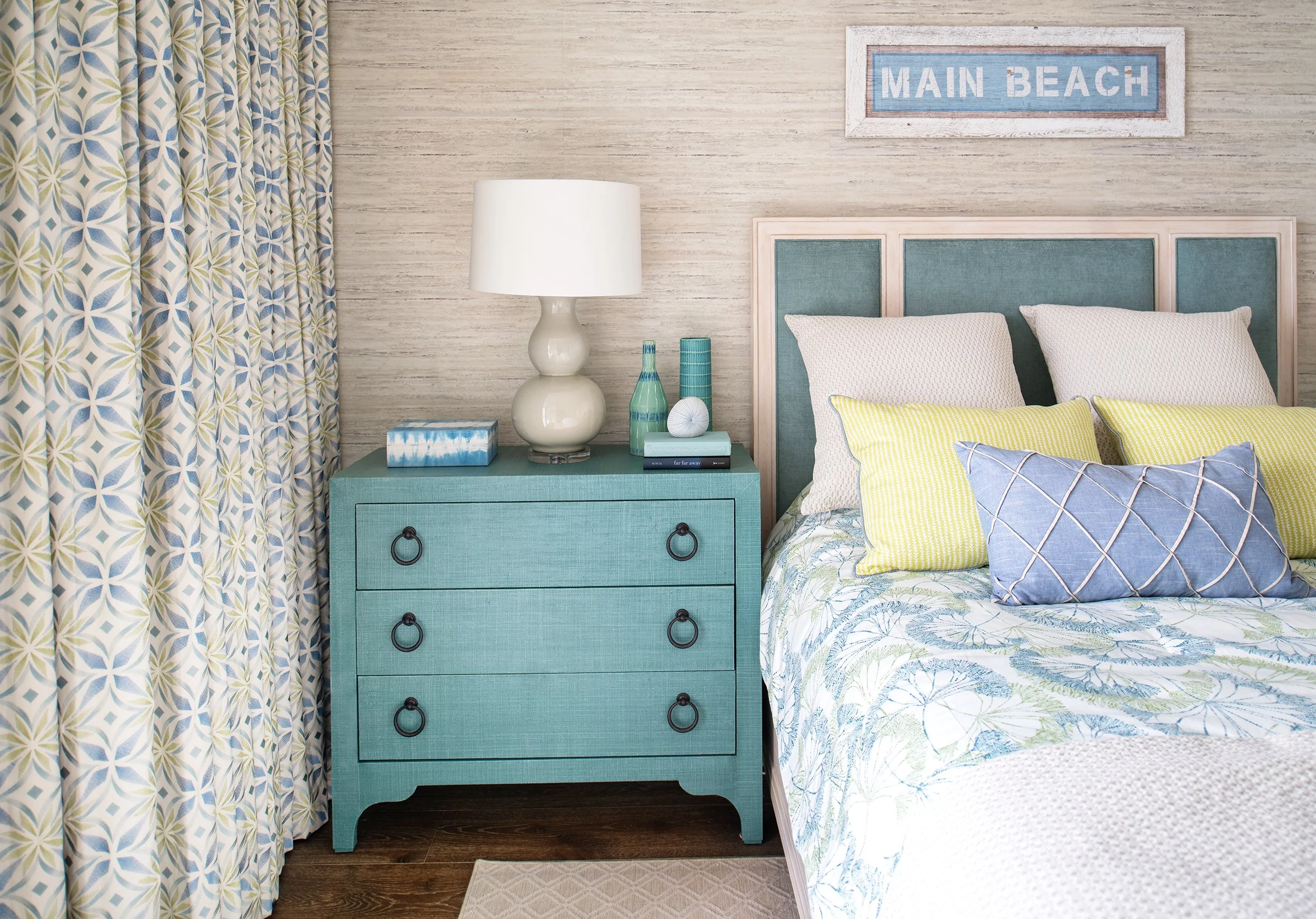 Beach-themed bedroom with a blue dresser, a white lampshade, decorative vases, and a bed with pastel yellow, blue, and white pillows, and a sign that says "Main Beach" above the bed.