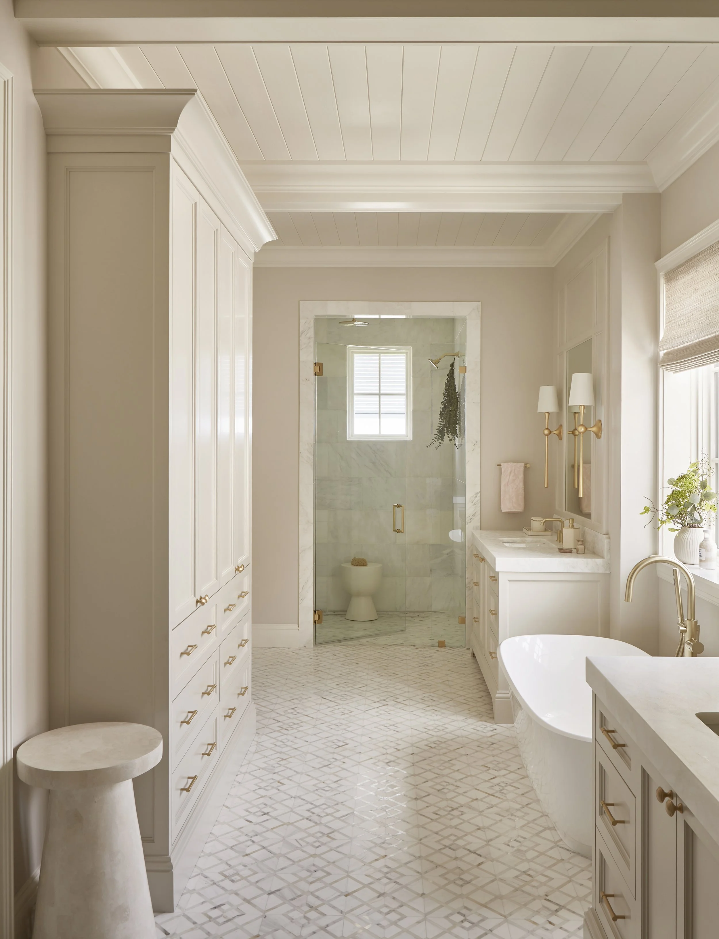 Bright and elegant bathroom with white cabinetry, a freestanding bathtub, a marble-topped vanity, and a glass-enclosed shower with a marble interior, window, and gold fixtures.
