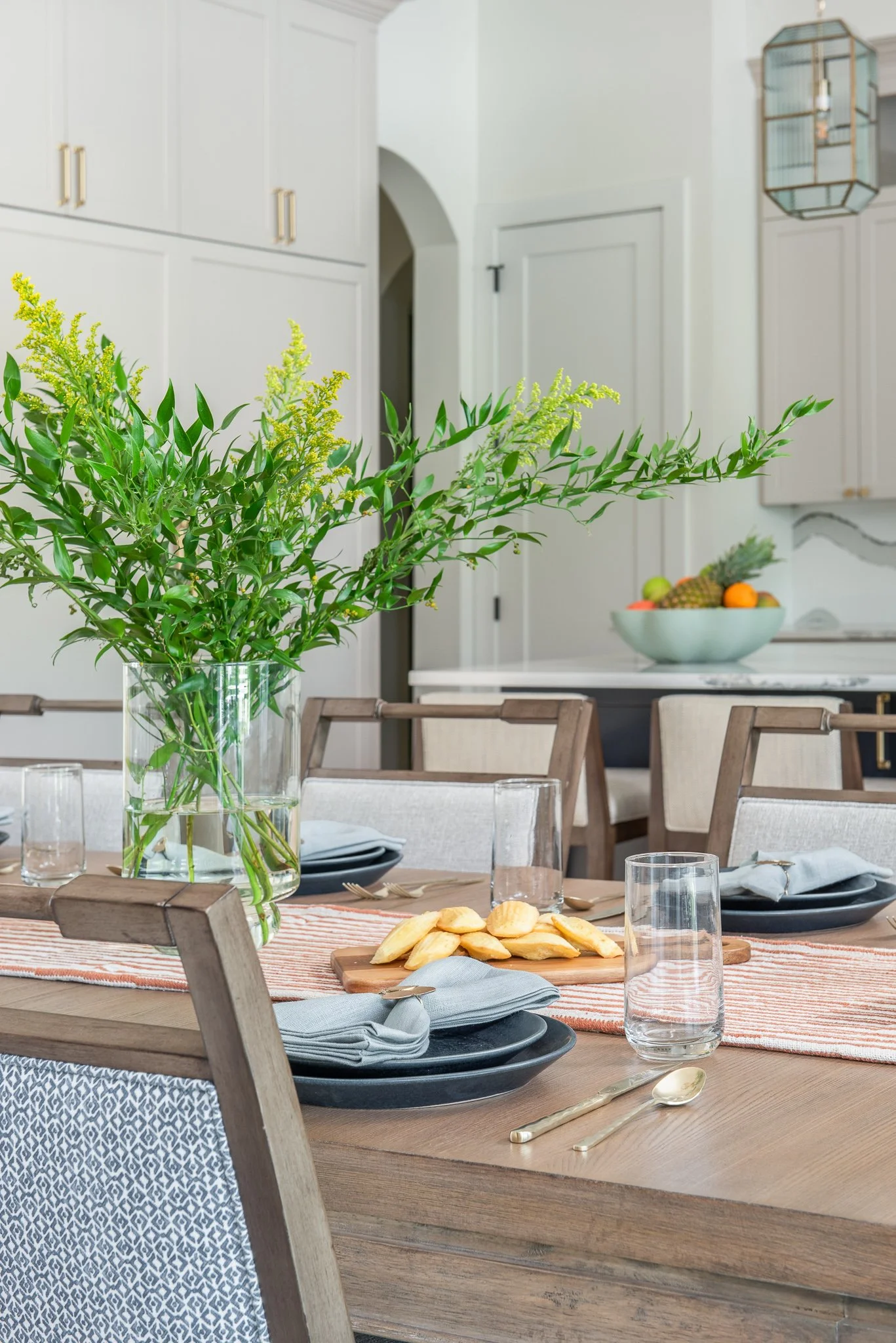 A dining table set with black plates, gold utensils, glasses, and napkins, with a large glass vase holding green leafy branches as a centerpiece, in a modern kitchen with white cabinets and a bowl of fruit in the background.