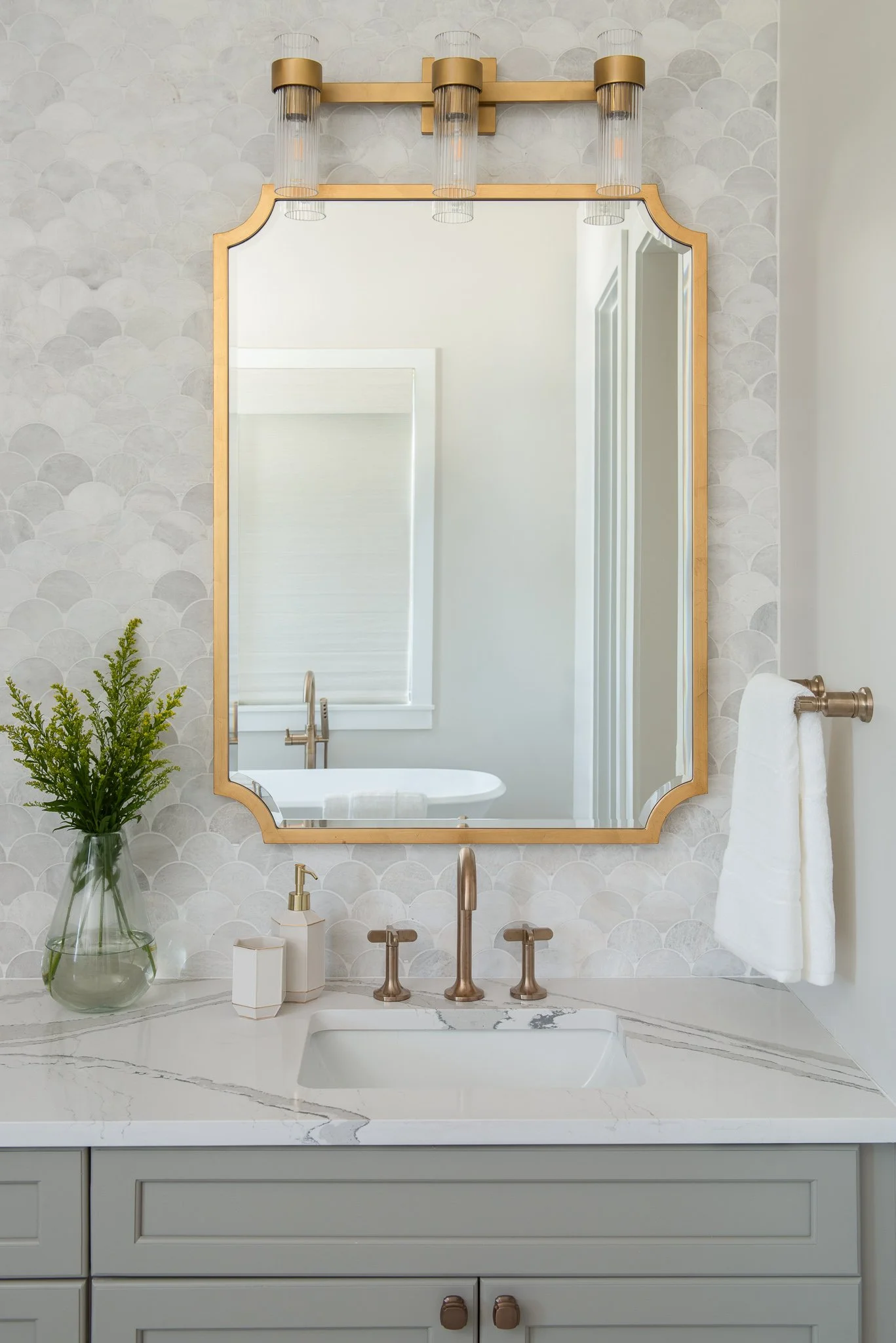Bathroom with a large mirror framed in gold, a white marble countertop with taupe-colored handles, a brass faucet, a clear glass vase with green plants, white soap and lotion dispensers, a white towel on a brass towel ring, and a gold light fixture w