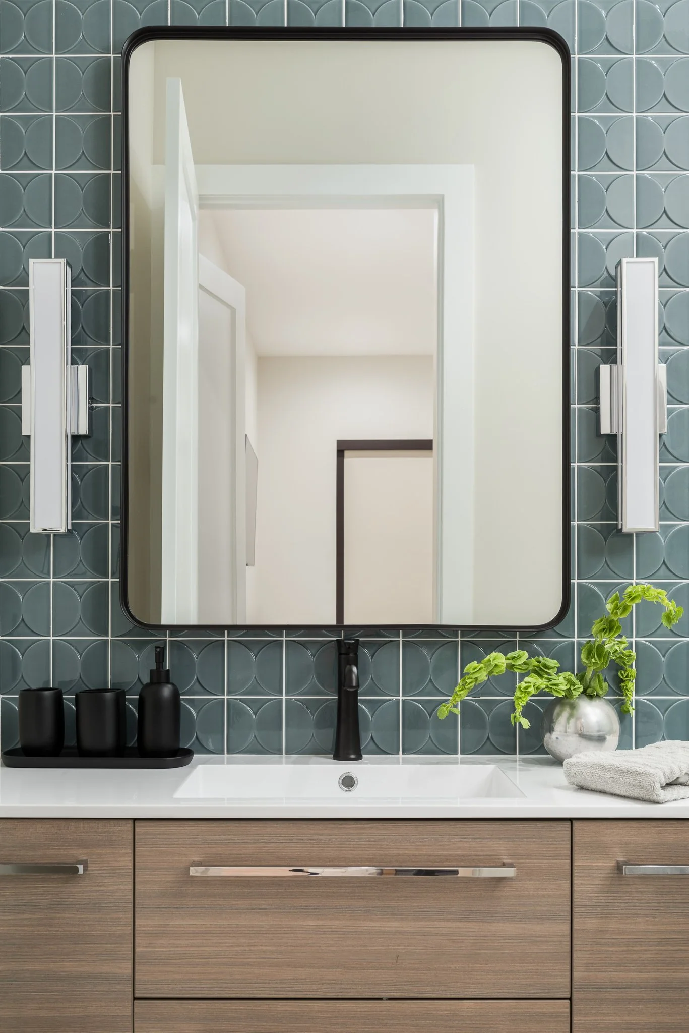 A bathroom vanity with a large rectangular mirror, black fixtures, and a plant in a silver vase. The wall behind the mirror is tiled with dark green tiles, and the vanity has a wood finish with a white countertop and a sink.