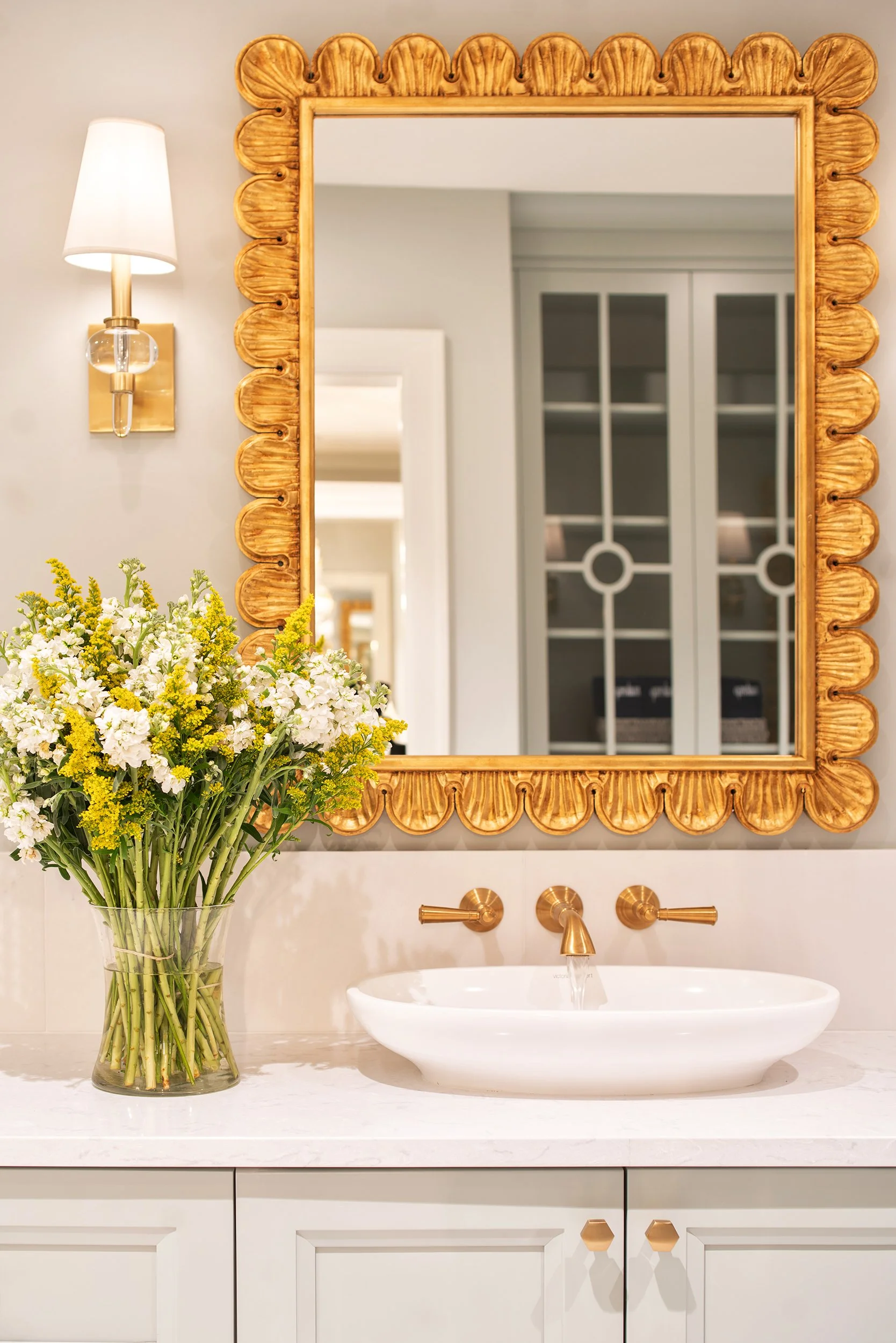 A bathroom vanity with a white countertop, a modern white vessel sink, gold fixtures, a large gold-framed mirror, a wall-mounted light, and a glass vase filled with white and yellow flowers.