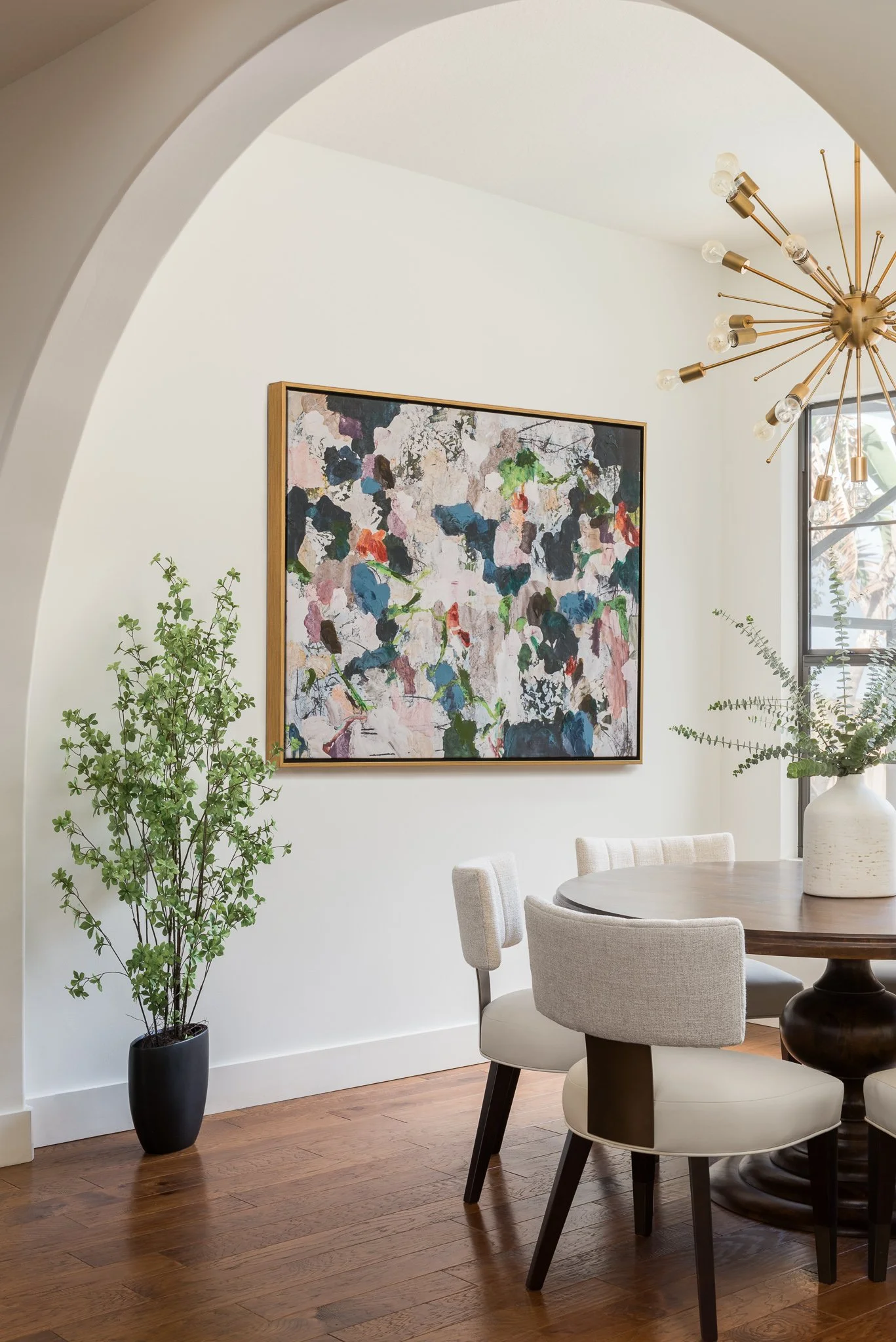 A dining room corner with a round dark wood table, four beige upholstered chairs, a white textured vase with green fern leaves, a large abstract painting, a black potted plant, a window, and a modern brass chandelier with multiple lights.