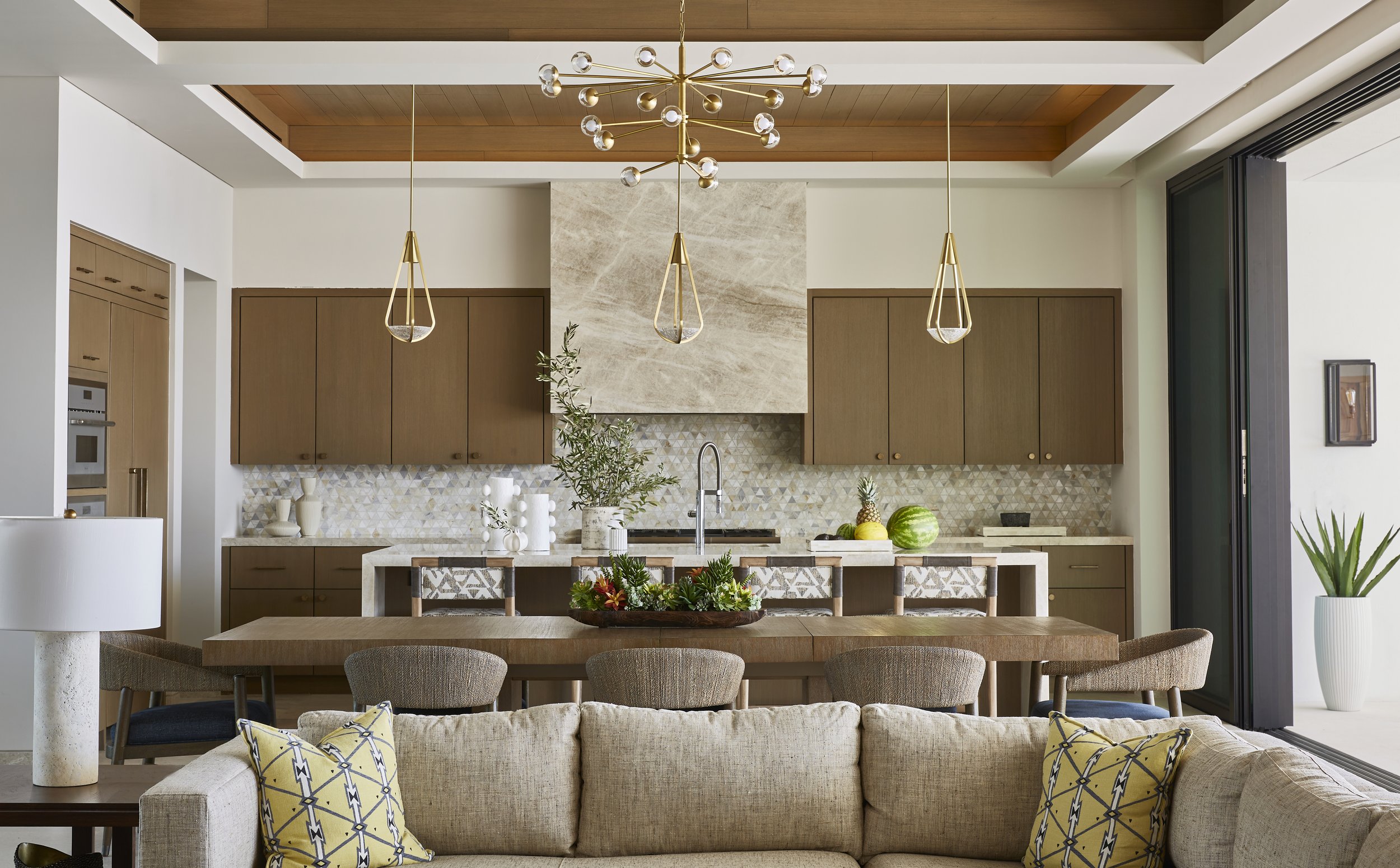 Modern open-plan kitchen and living room with a beige sectional sofa, yellow patterned pillows, a wooden dining table, pendant lights, and kitchen cabinets with a marble backsplash.