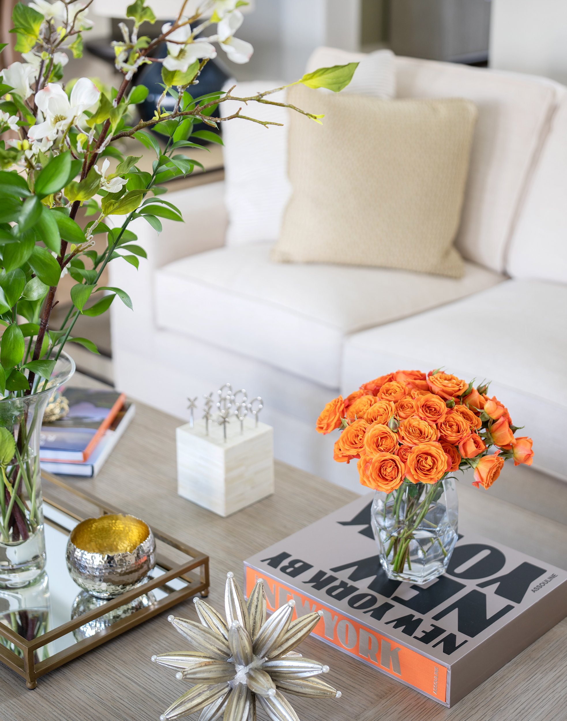 Living room with a coffee table decorated with flowers, books, and decorative objects, including a large arrangement of orange roses in a glass vase.