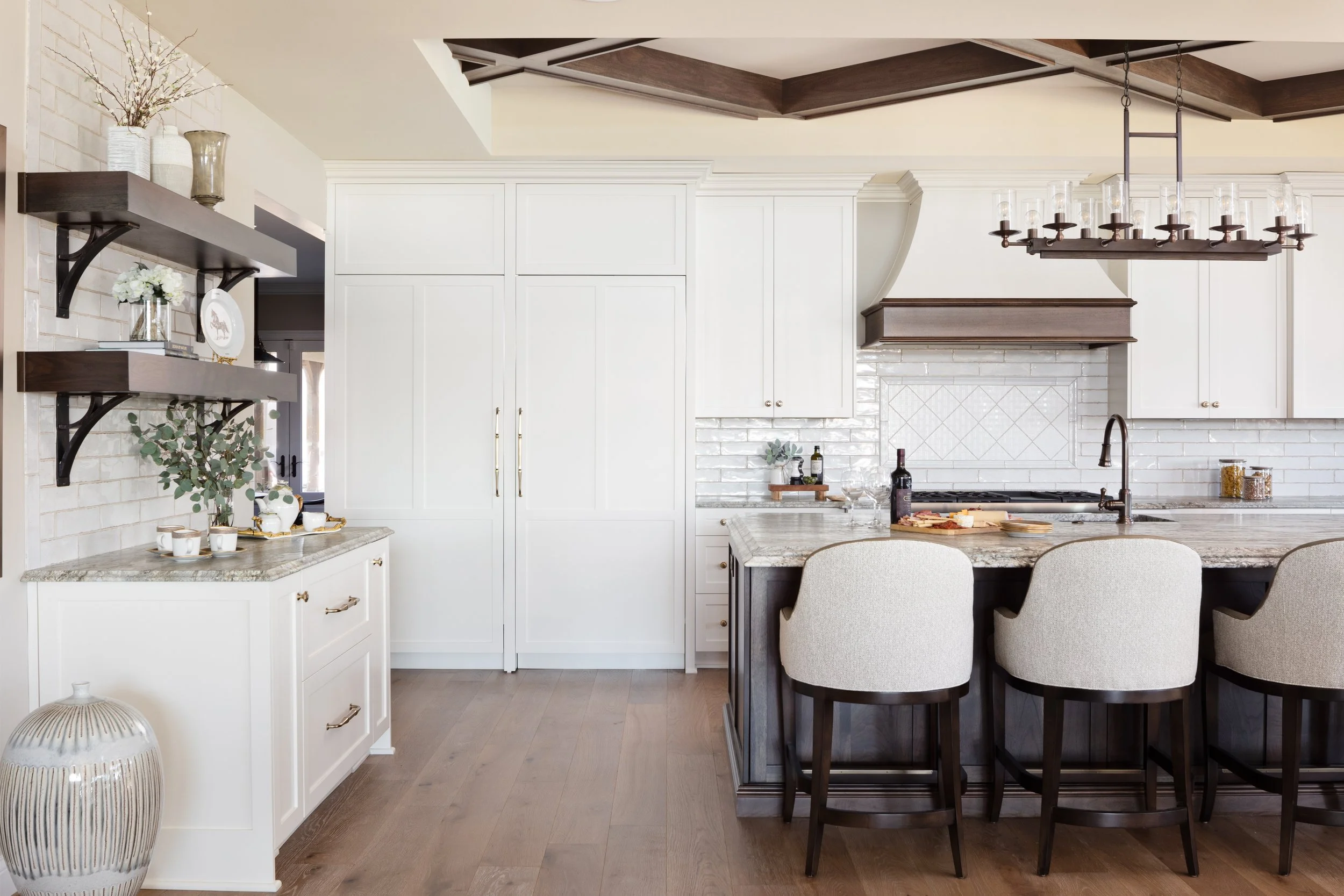Bright kitchen with white cabinets, a marble island with bar stools, dark wood accents, and decorative shelves on the wall.