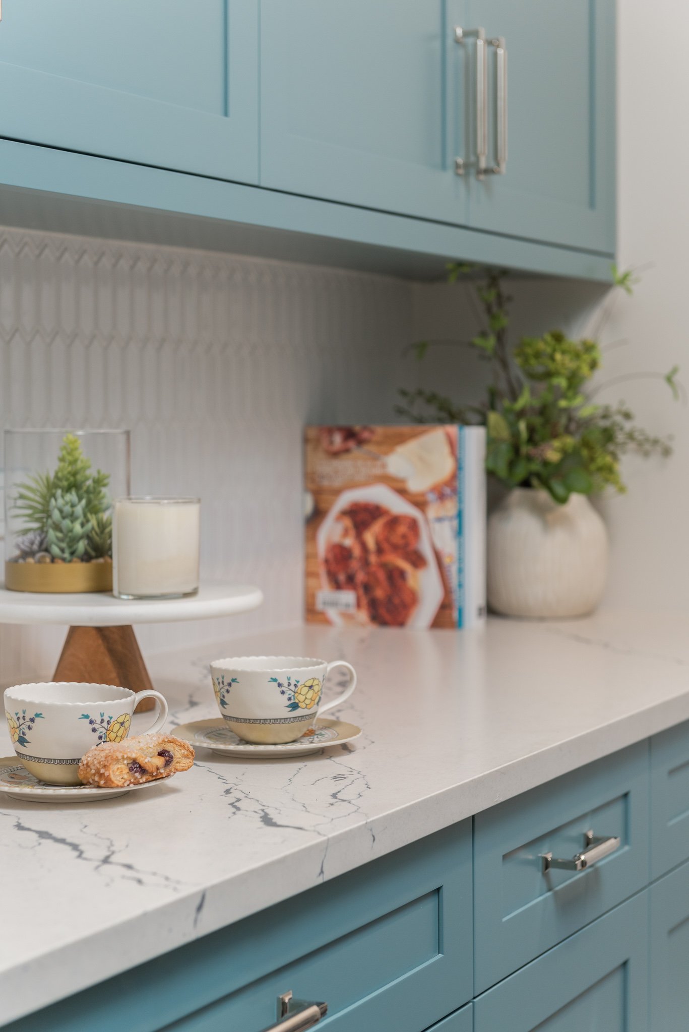 A kitchen countertop with two cups of tea, a cookie, a potted succulent, a white candle, and a cookbook, with blue cabinetry and a potted plant in the background.