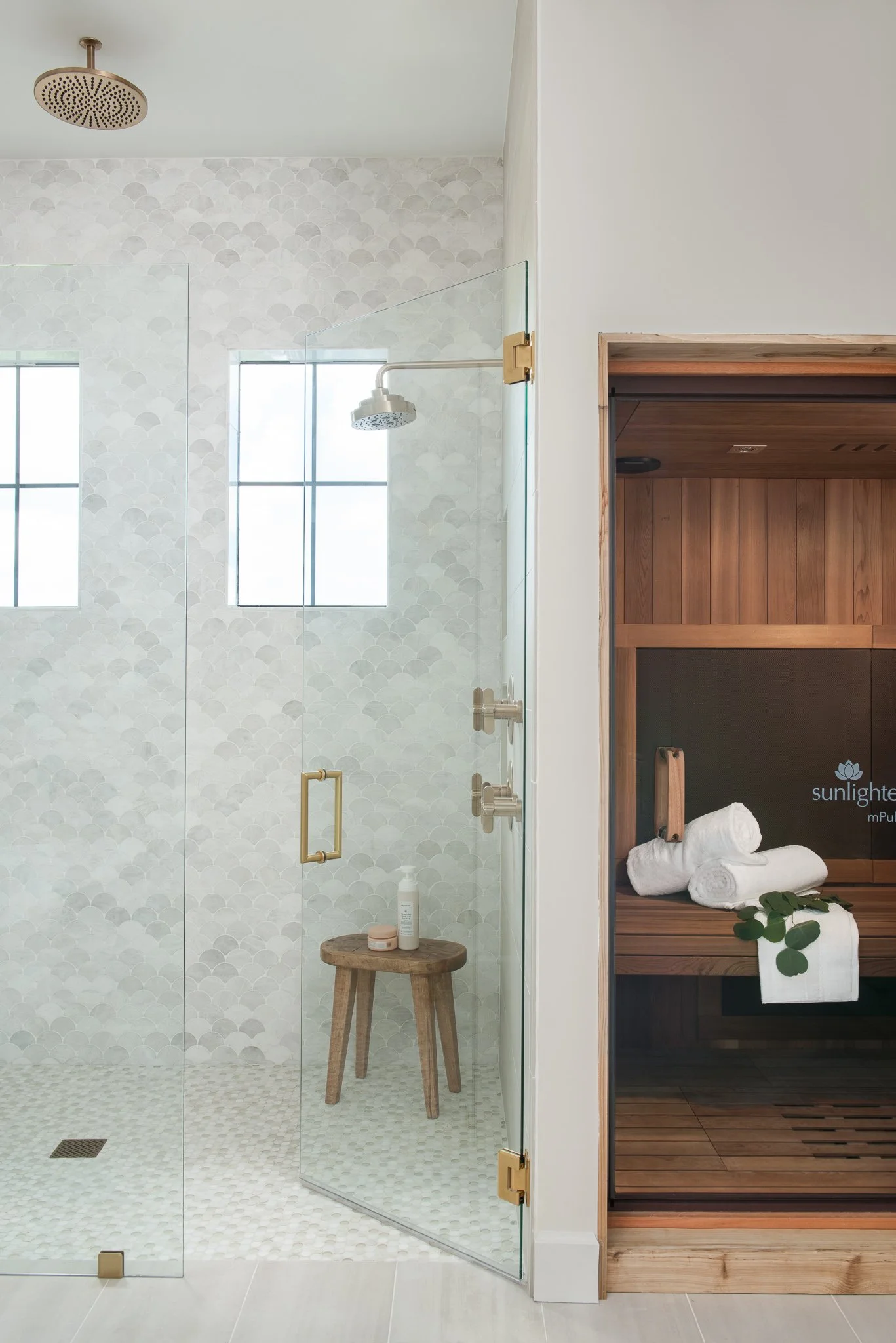 Modern bathroom with glass shower enclosure, a wooden stool inside the shower holding toiletries, and a sauna room with rolled white towels and green leaves outside.
