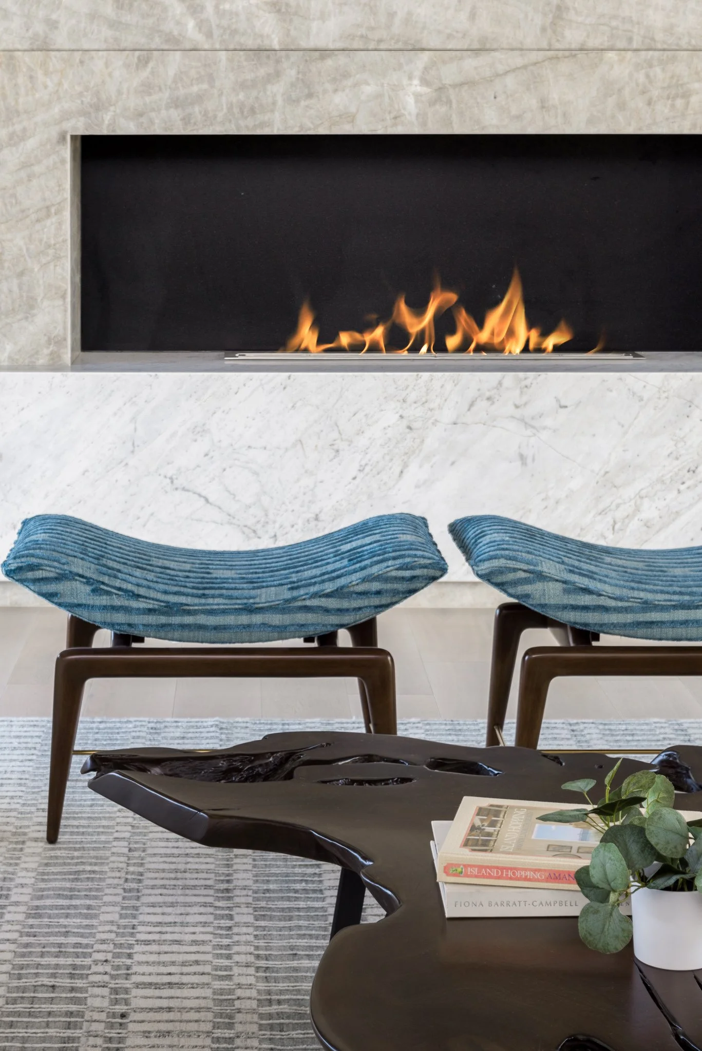 A modern living room with a fireplace, blue velvet stools, a black side table with books, and a potted plant.