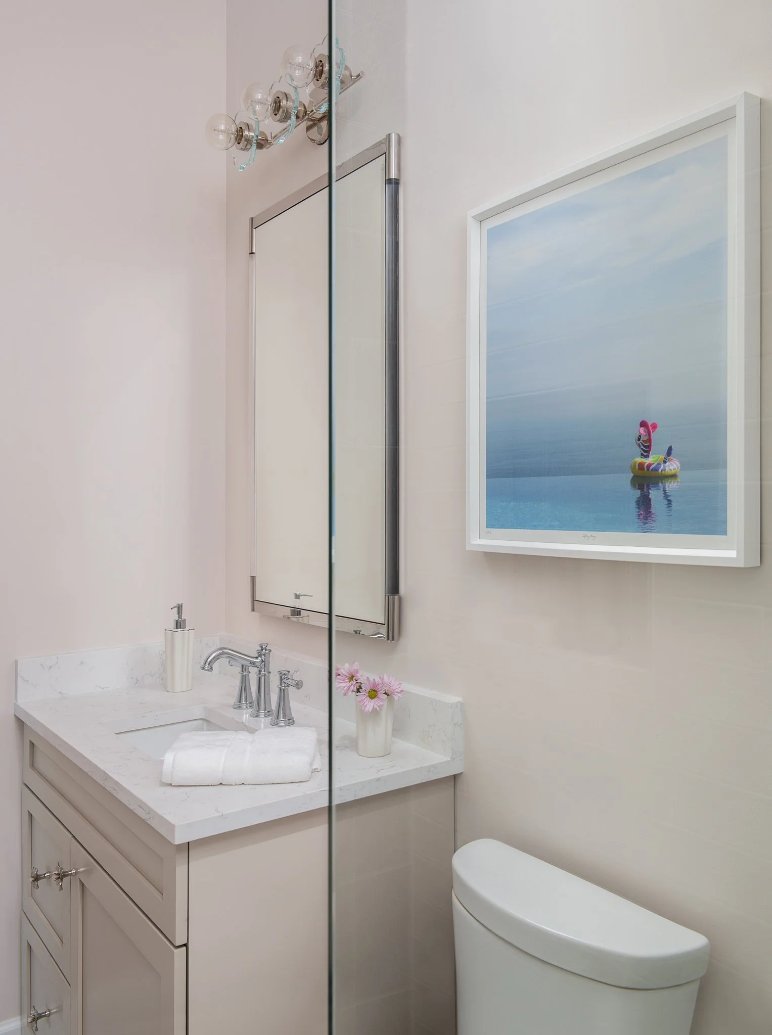 A bathroom with a white vanity sink, a soap dispenser, pink flowers in a white vase, a mirror, a framed picture of a cartoon character in a pool, and part of a toilet.