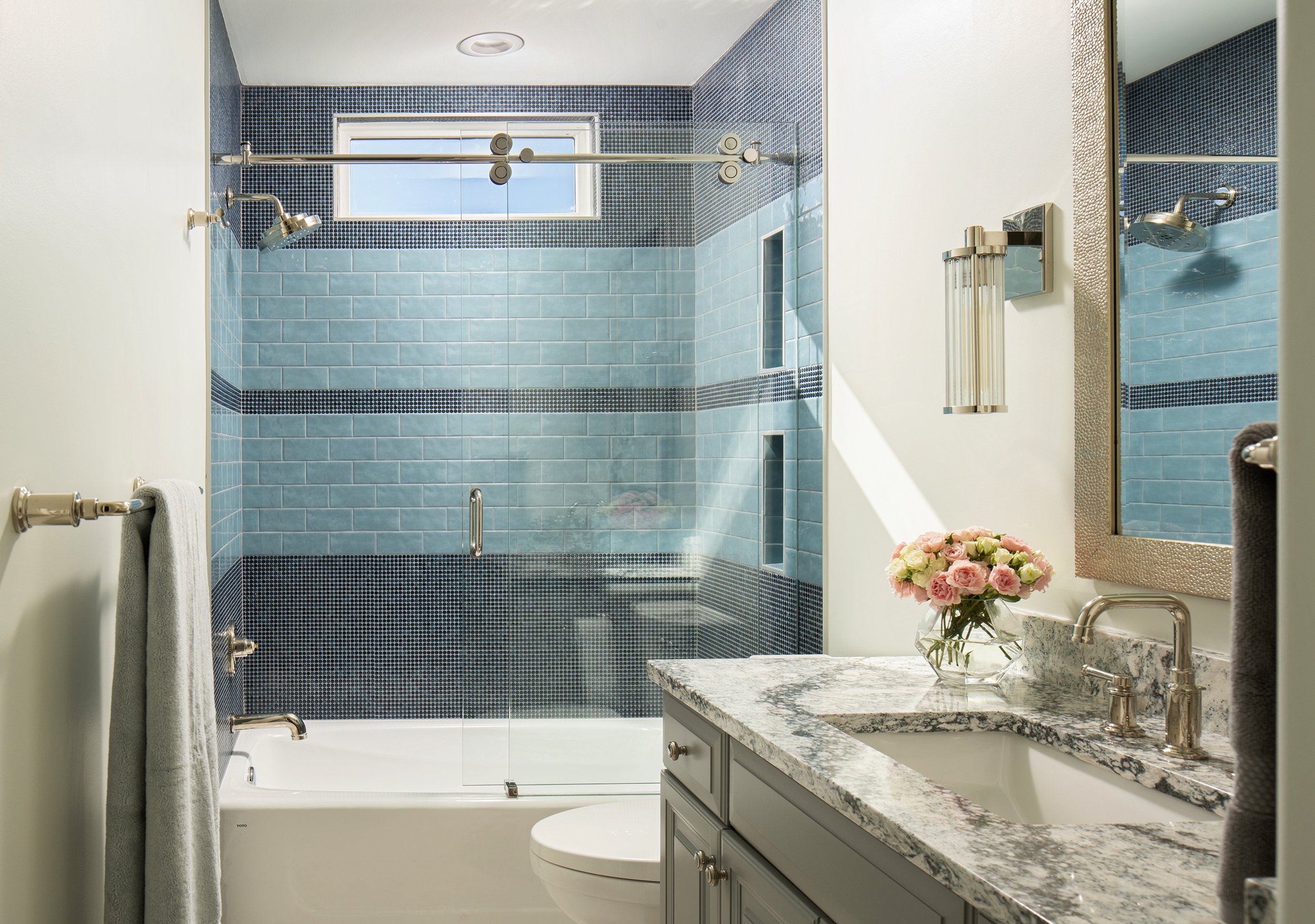 Modern bathroom with a blue tiled shower, a white bathtub, a gray vanity with a marble countertop, a round mirror, a bouquet of pink roses, and a window providing natural light.