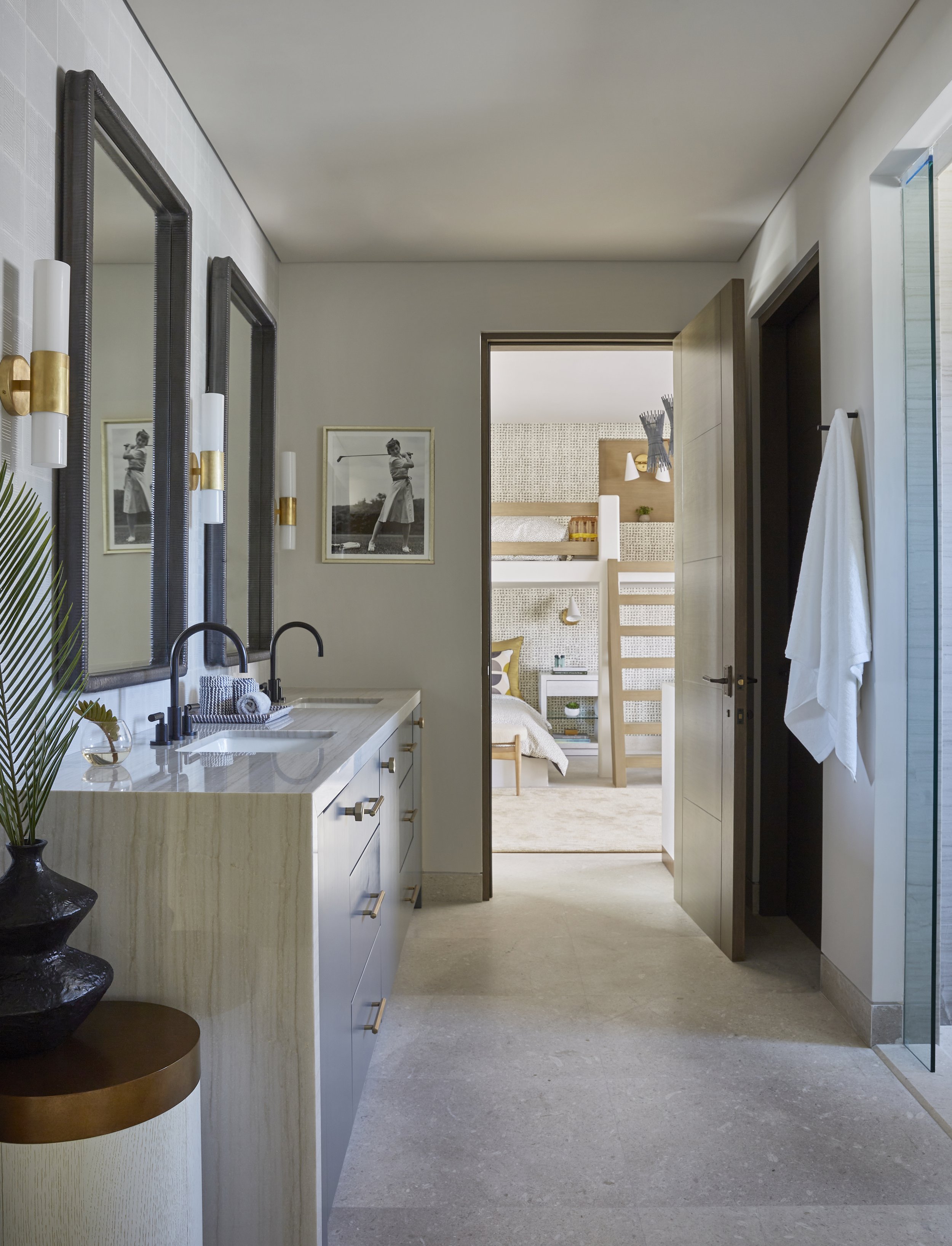 A modern bathroom with white cabinetry and a marble countertop, gold hardware, and three framed black-and-white photos on the wall. There is a large mirror, a black faucet, and a potted plant on a small table. Through an open door, a bedroom with bun