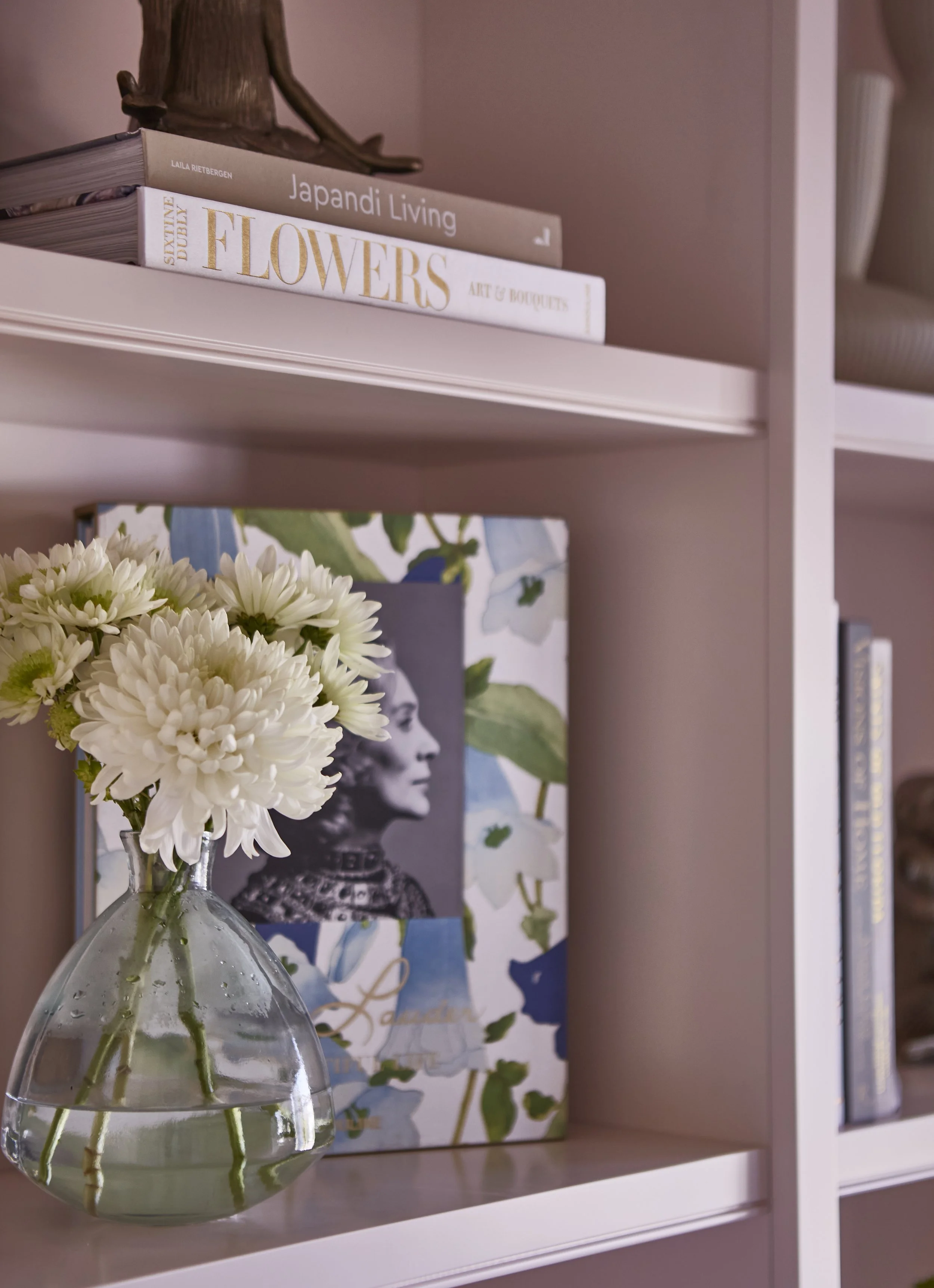 A white bookshelf with books titled 'Japandi Living', 'Flowers', and other books, along with a glass vase containing white flowers, and framed photographs of portraits and nature.