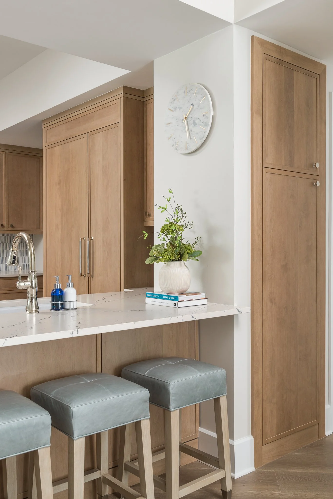 A kitchen with wooden cabinets, a white marble counter, a potted plant on top of the counter, and three gray padded stools. A white wall with a clock is visible.