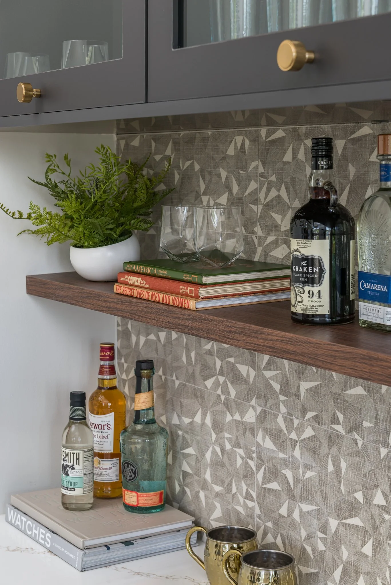 A kitchen counter with a patterned backsplash, a wooden shelf holding a white pot with green fern, three stacked books, two glass tumblers, and bottles of alcohol. There are also liquor bottles on a lower surface, a white book titled 'Watches,' and t