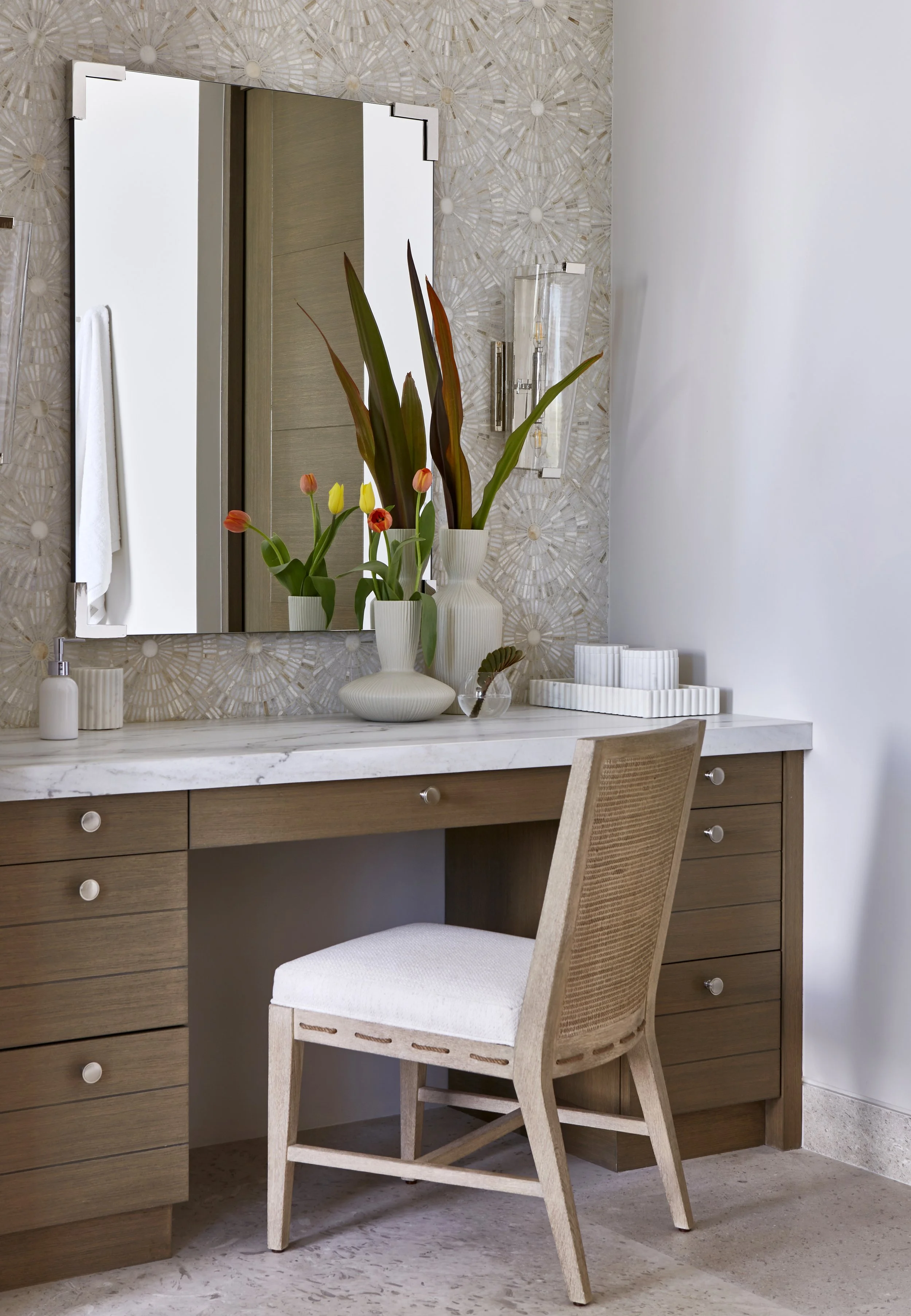 A bedroom vanity area with a wooden desk, a beige upholstered chair, a large wall mirror, and decorative vases with flowers. The wall behind the mirror has a textured geometric pattern.