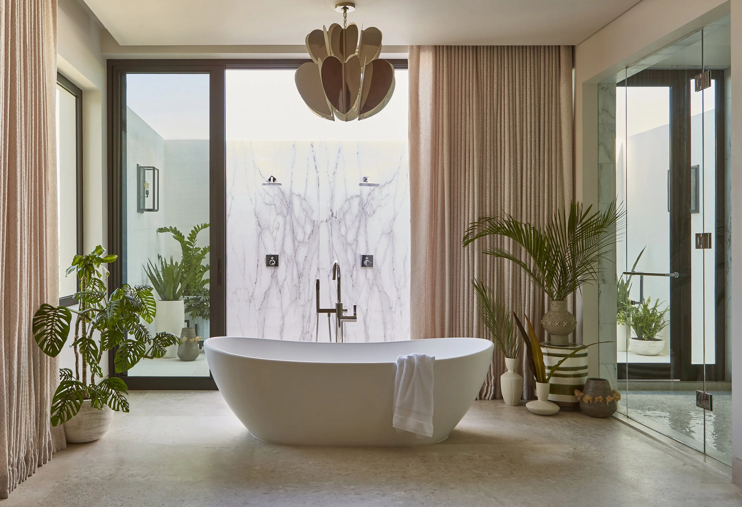 Modern bathroom with freestanding bathtub, large windows, indoor plants, marble accent wall, and glass shower enclosure.