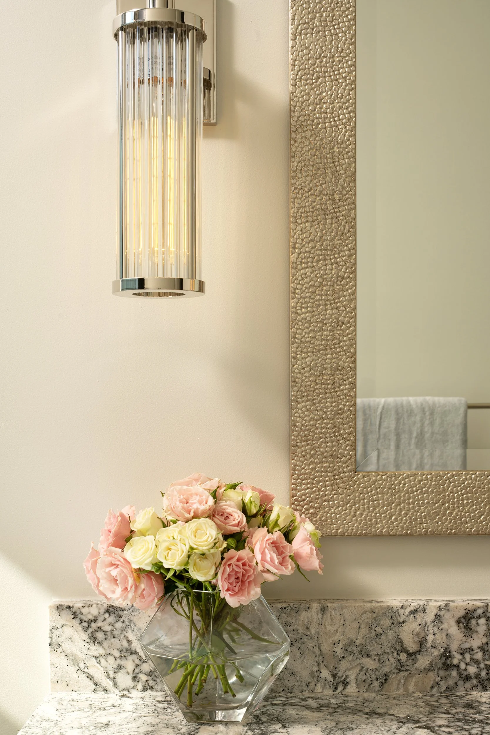 A decorative wall sconce light fixture, a large textured silver-framed mirror, and a glass vase with pink and white roses on a granite countertop.