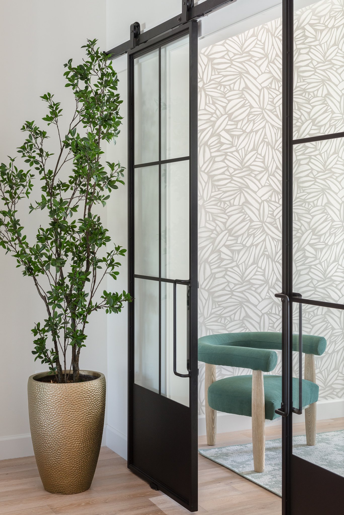 Interior view of a room with a large potted green plant, a black-framed glass door, a patterned wall, a green upholstered chair, and light wood flooring.