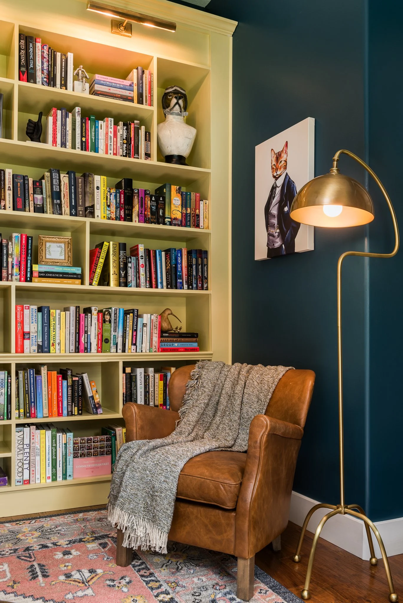 A cozy reading nook with a tan leather armchair draped with a gray throw blanket, a colorful patterned rug, a white bookshelf filled with books and decorative items, a gold floor lamp with a curved arm and round shade, and a portrait of an anthropomo