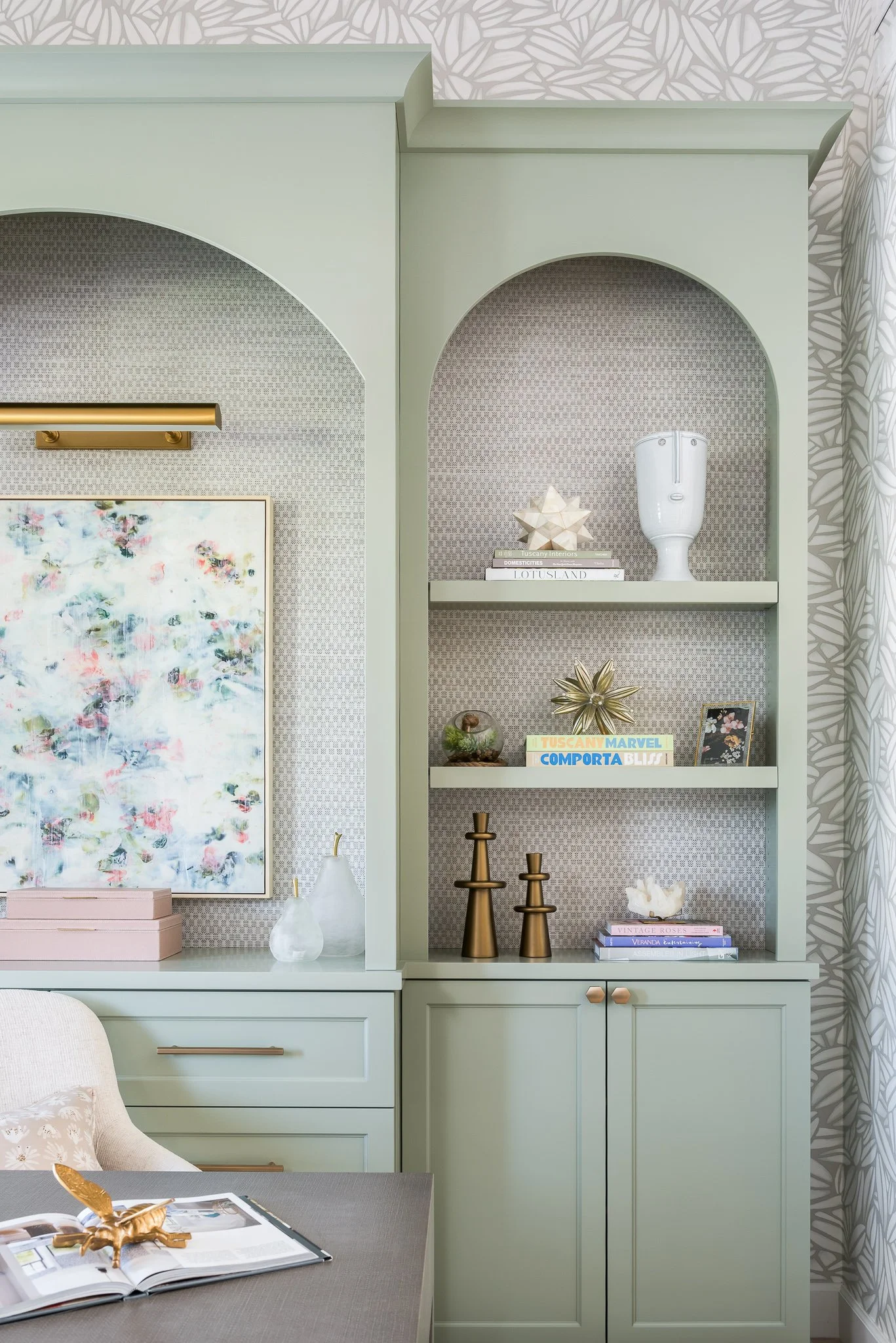 A mint green built-in cabinet with open shelves and closed storage below, decorated with books, vases, figurines, and framed art, against patterned wallpaper and a leafy curtain.