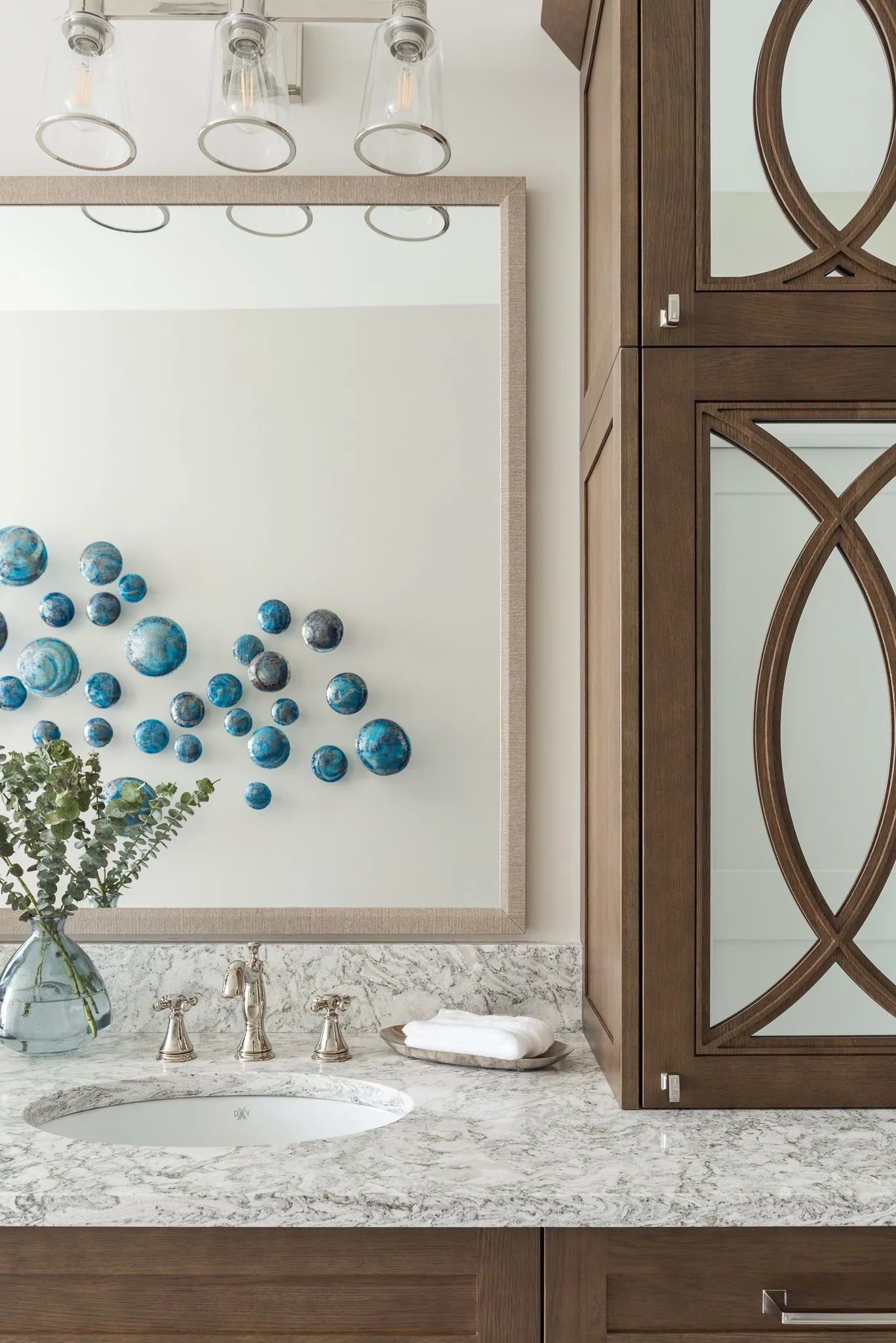 Bathroom vanity with a marble countertop, small oval sink, silver fixtures, a clear vase with green foliage, and a large mirror. Decor includes a cluster of blue spherical wall art and a wooden cabinet with mirrored doors.