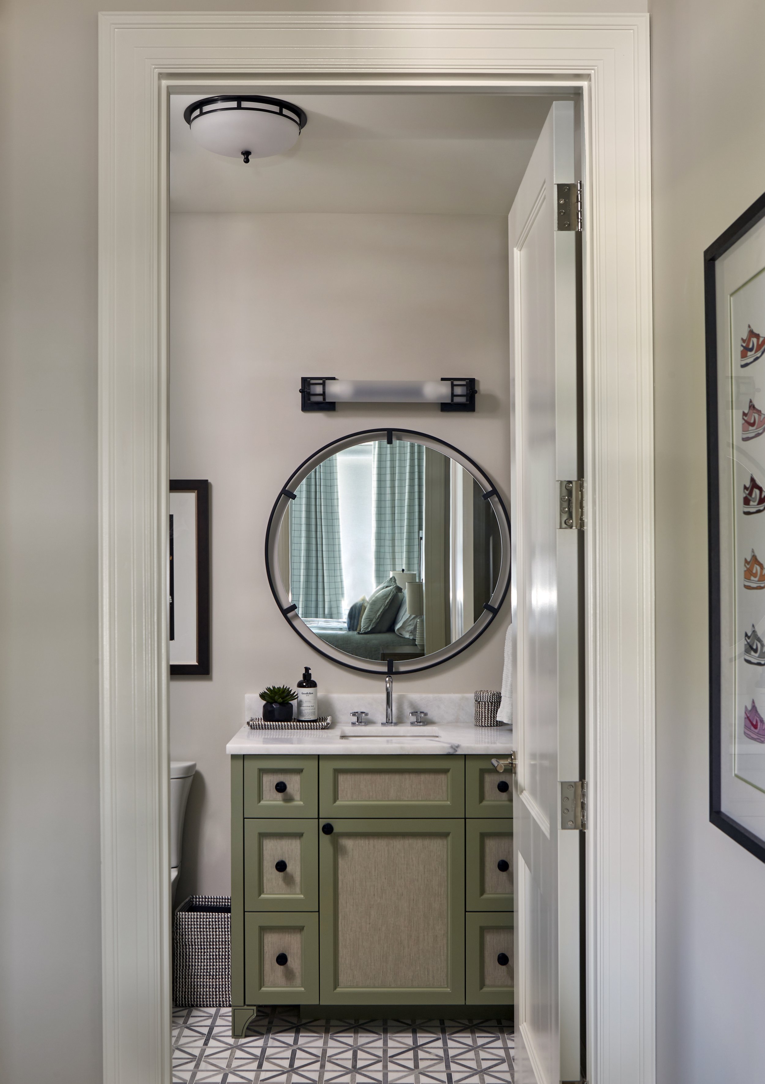 Bathroom with a green vanity, marble countertop, round mirror, small potted plant, soap dispenser, and framed artwork on the wall.