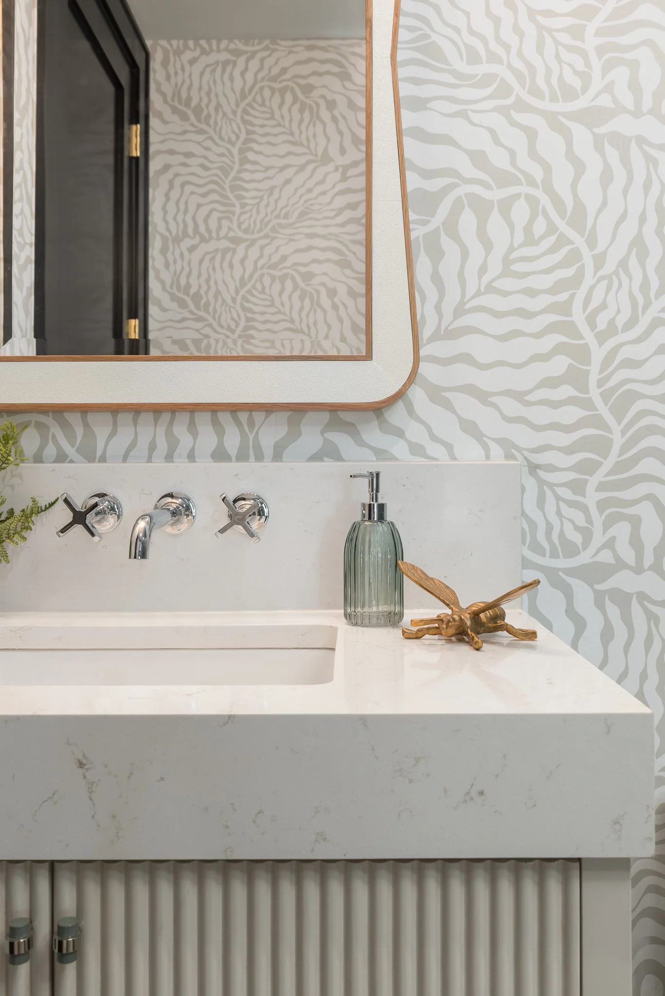 A bathroom vanity with a white marble countertop, a green soap dispenser, a gold insect sculpture, a mirror, and chrome fixtures, set against wallpaper with a white and gray leaf pattern.
