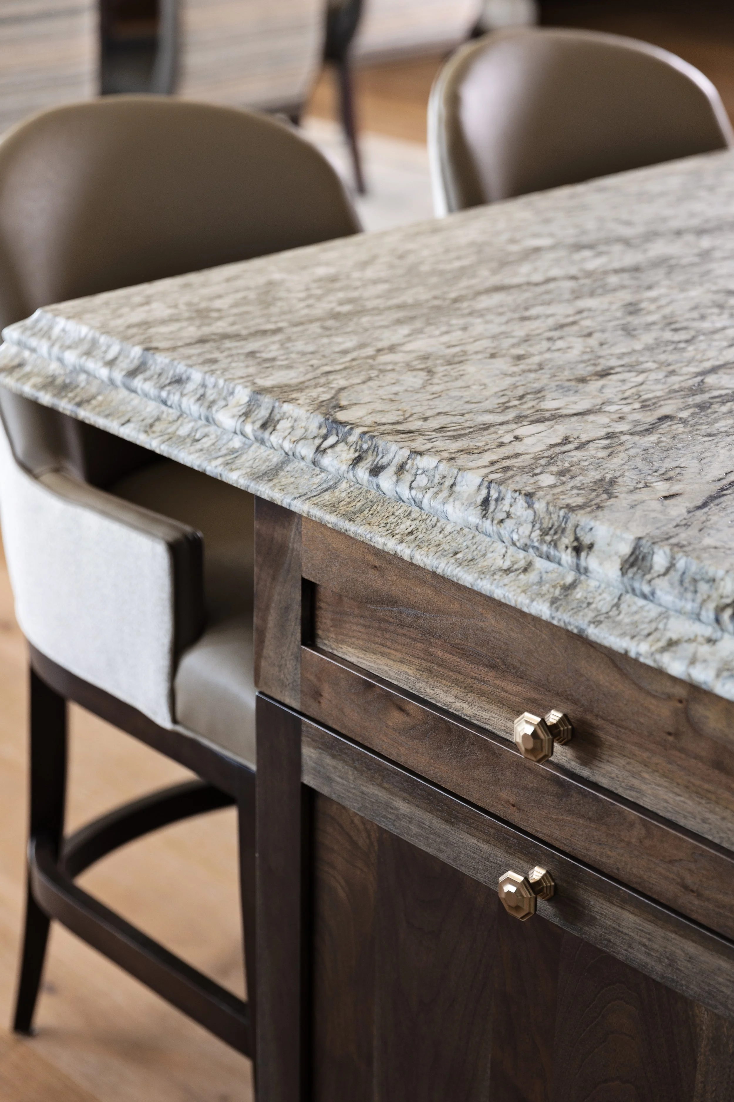 Close-up of a kitchen island with a polished granite countertop and dark wood cabinets with gold knobs, surrounded by beige upholstered barstools.