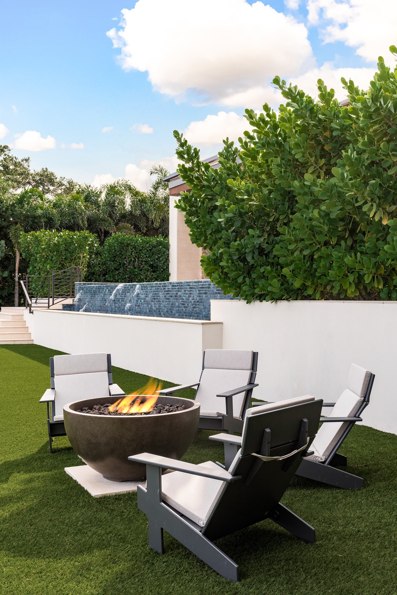 Backyard patio area with four outdoor chairs surrounding a firepit. The yard features a grassy lawn, a white wall, a small waterfall, and lush green trees and shrubs under a partly cloudy sky.
