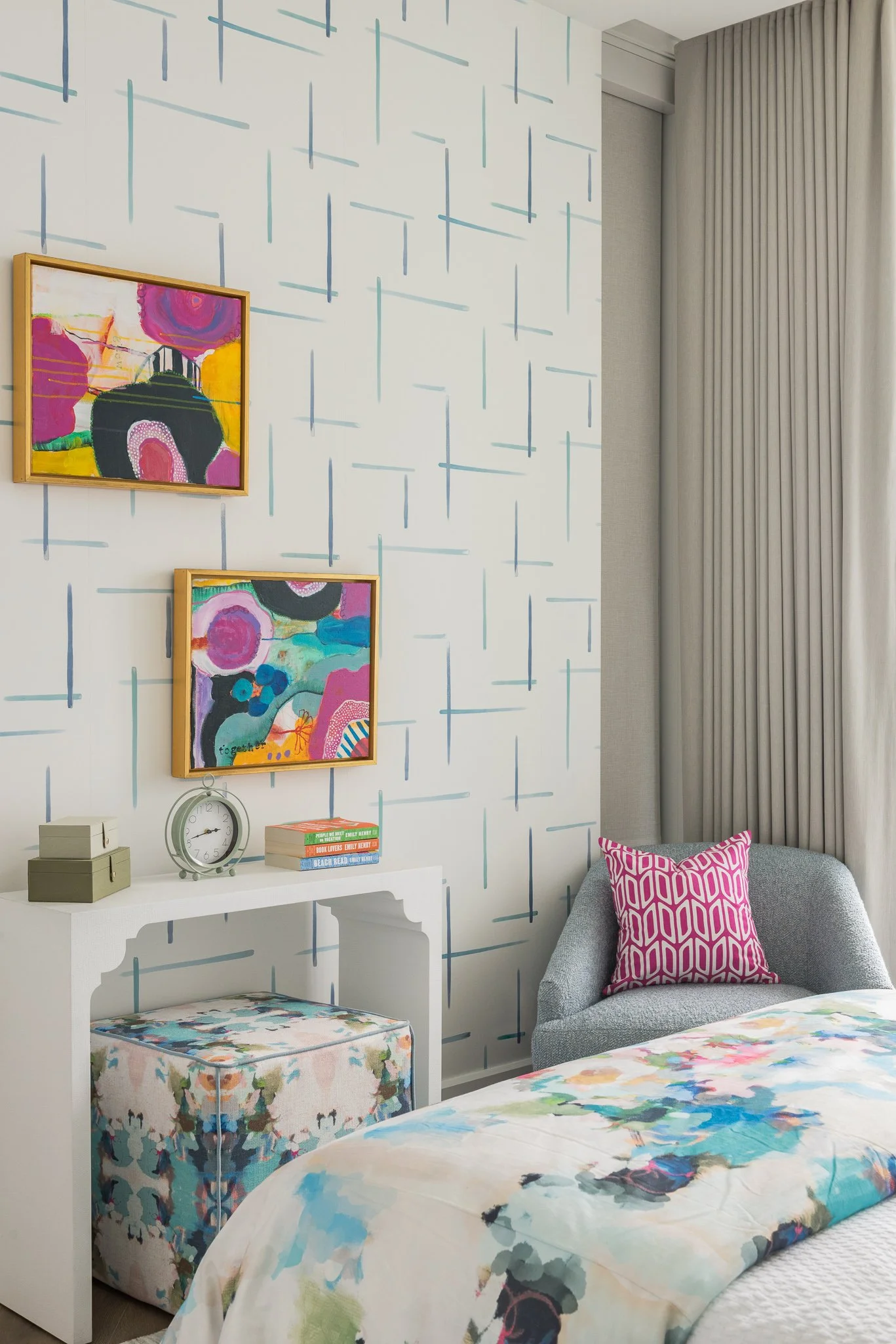 A bedroom corner with a bed, a gray armchair with a pink patterned pillow, a white desk with books, a clock, and storage boxes, decorated with abstract wall art and patterned curtains.
