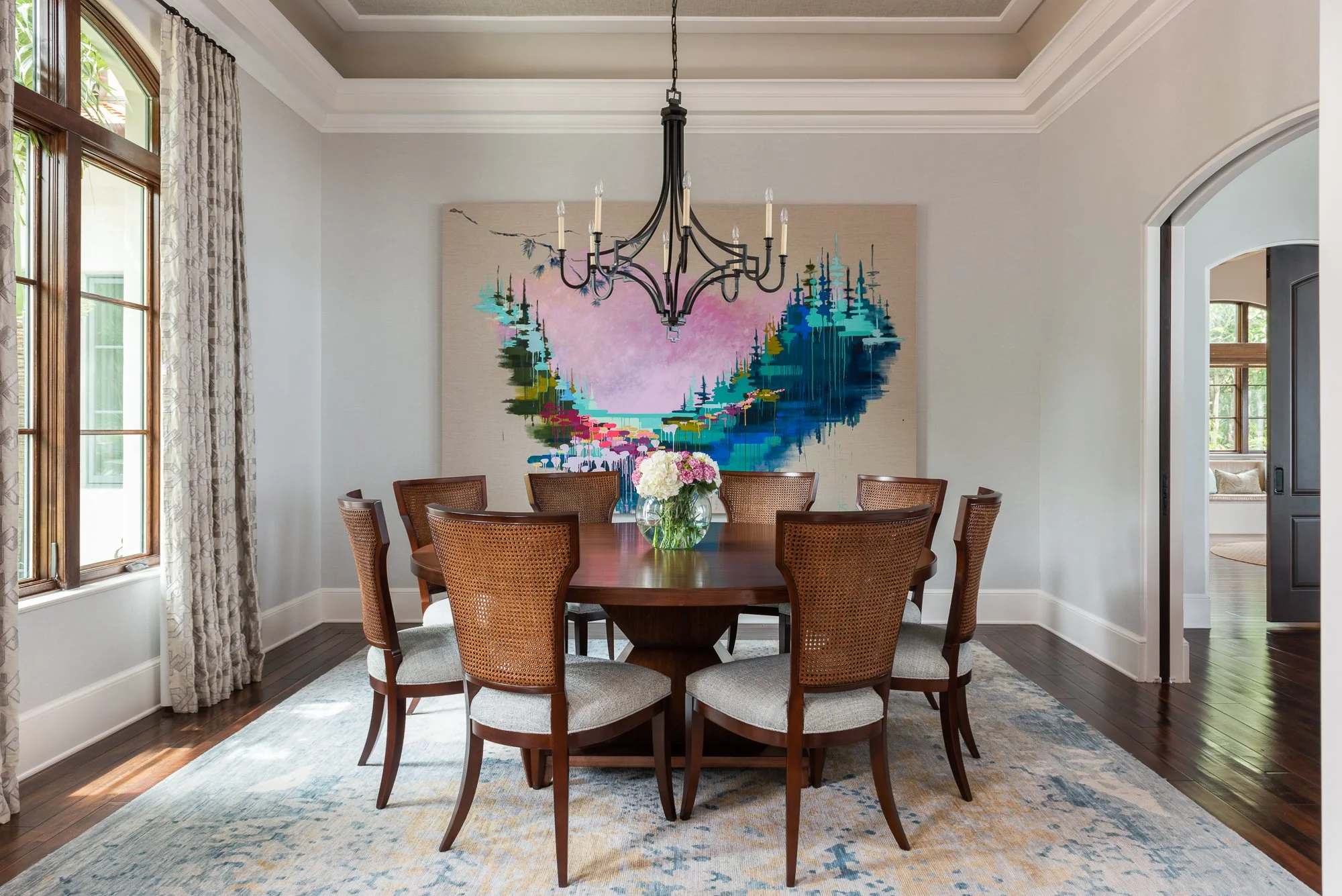 A dining room with a wooden table and eight matching chairs with woven backs, a glass vase centered on the table with white and pink flowers, a chandelier above, a colorful landscape painting on the wall, and windows with patterned curtains.