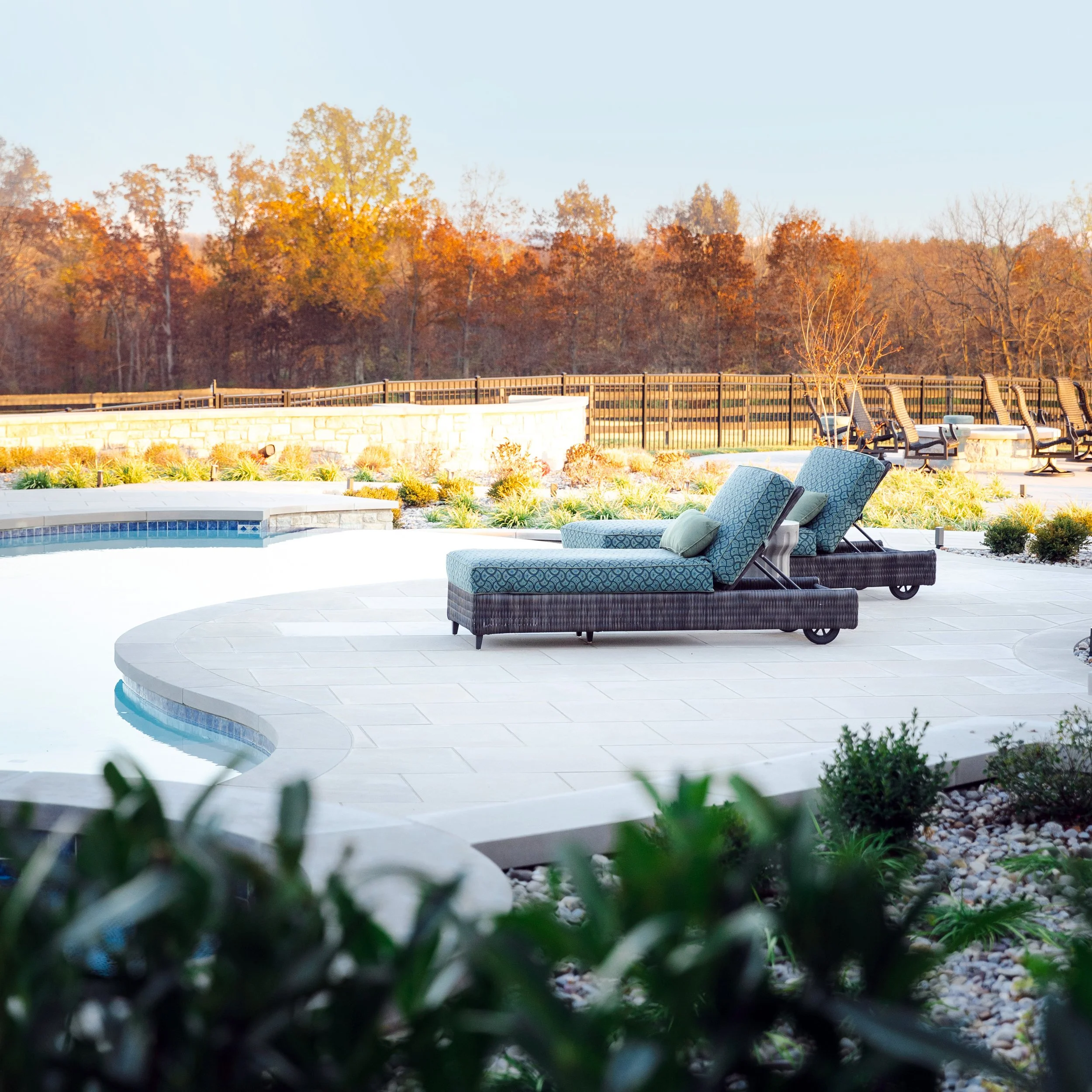 Poolside scene with two lounge chairs and pillows, surrounded by a landscaped garden and trees with autumn foliage.