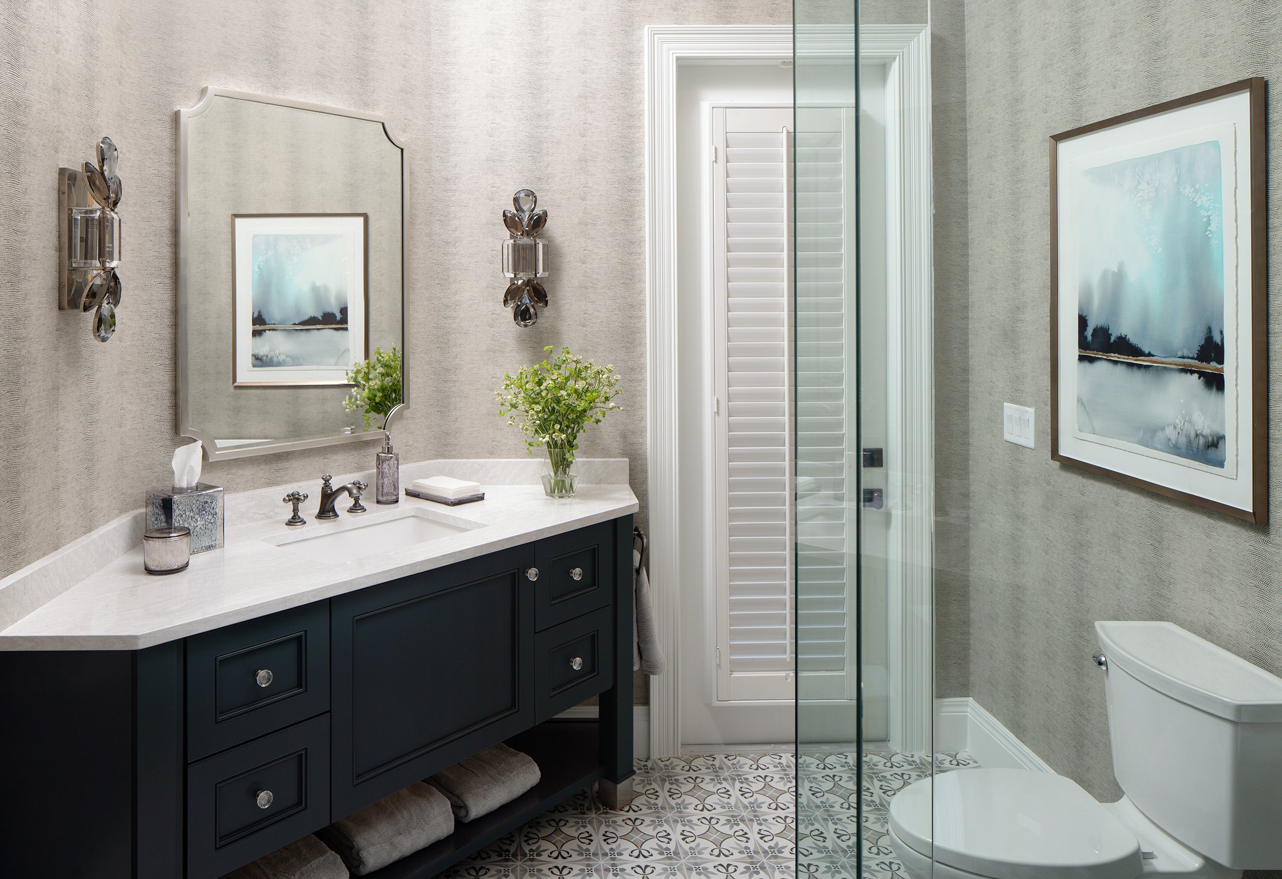 A modern bathroom with a navy blue vanity with a white marble top, a large mirror, artwork on the walls, a glass-enclosed shower, and a white toilet. There are decorative lighting fixtures and green plants for decor.