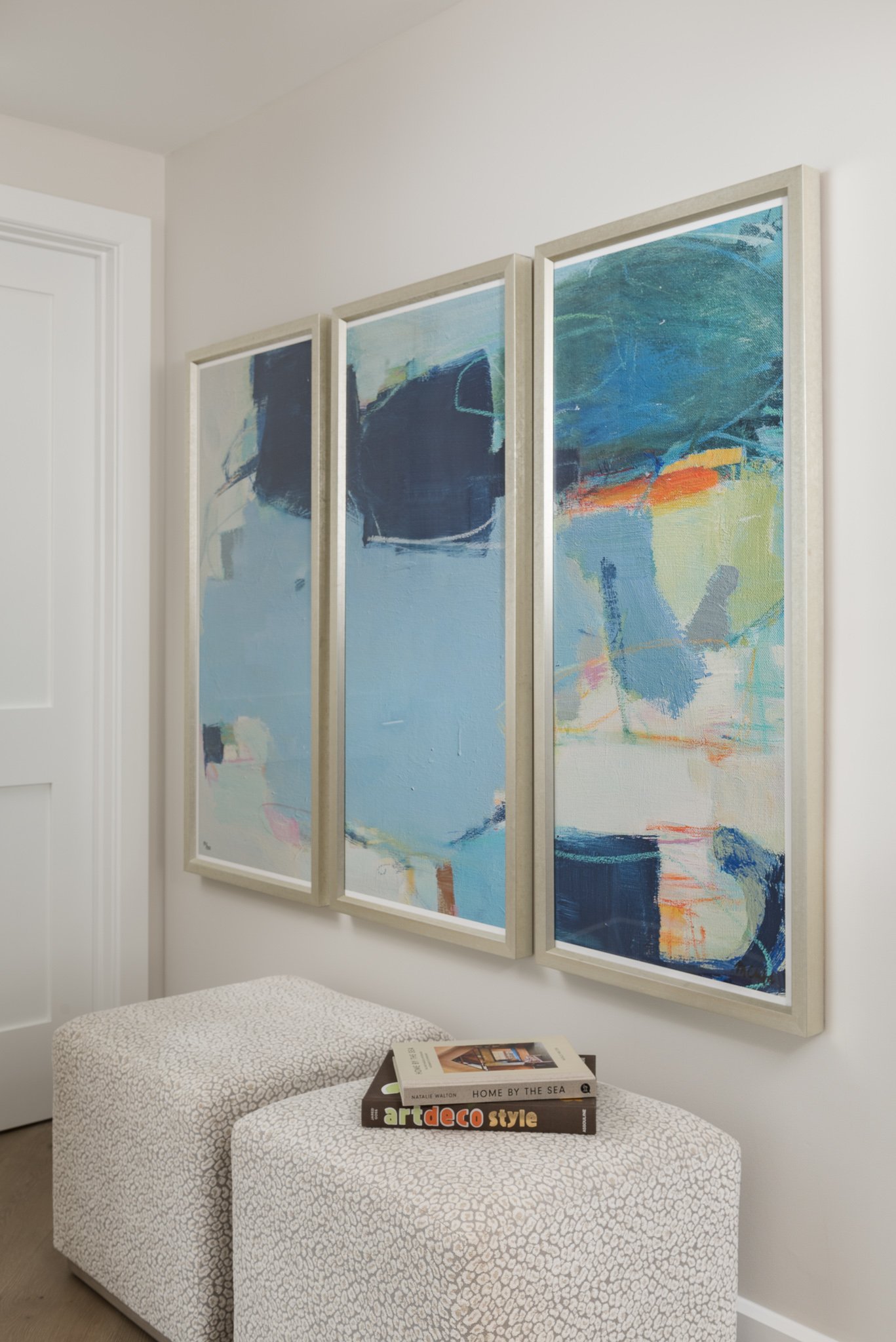 Three abstract paintings in white frames hanging above a pair of upholstered ottomans with books on top in a bright white room.