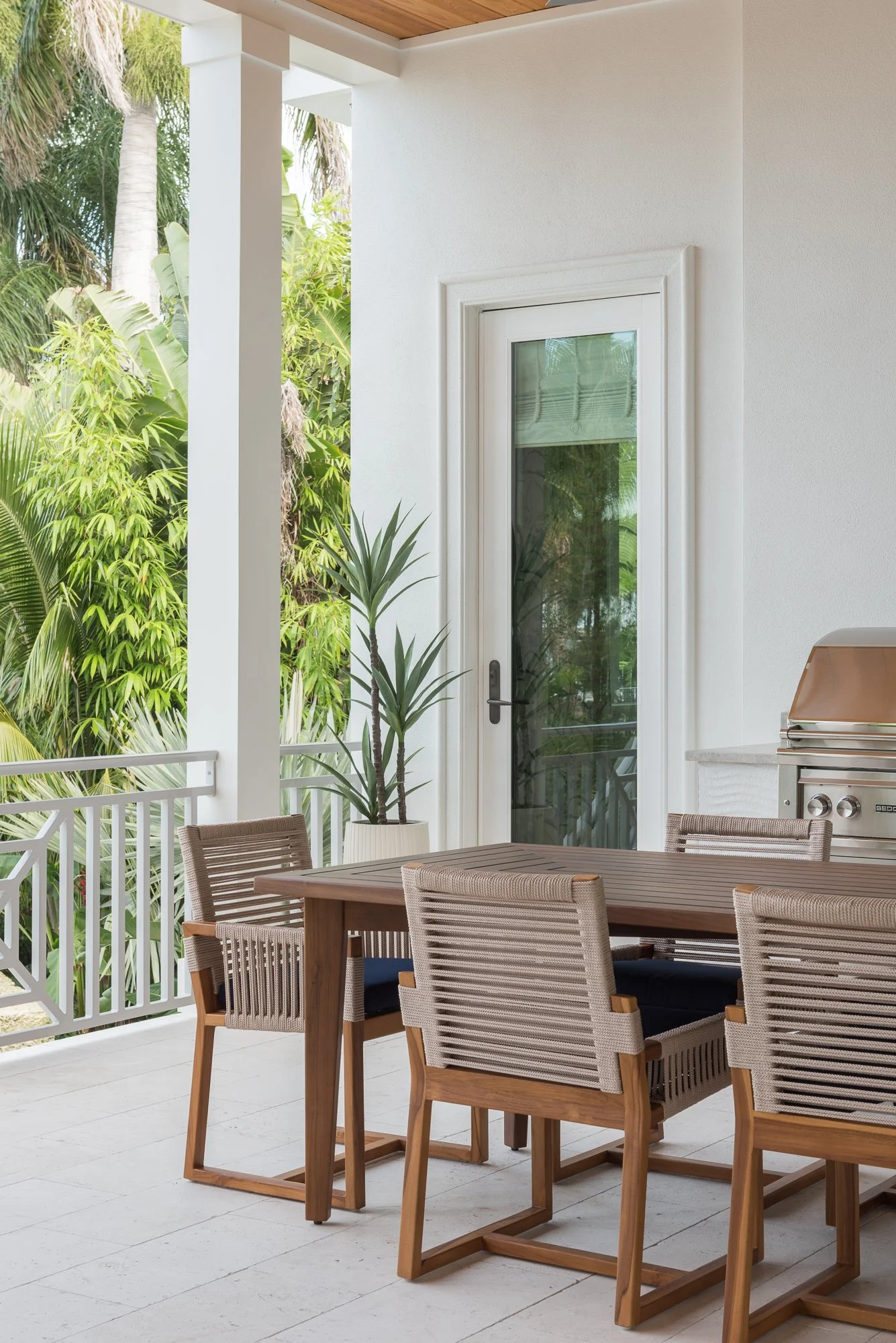 An outdoor patio with a wooden dining table, six matching chairs, a potted plant, a white railing, a glass door, and lush green trees in the background.