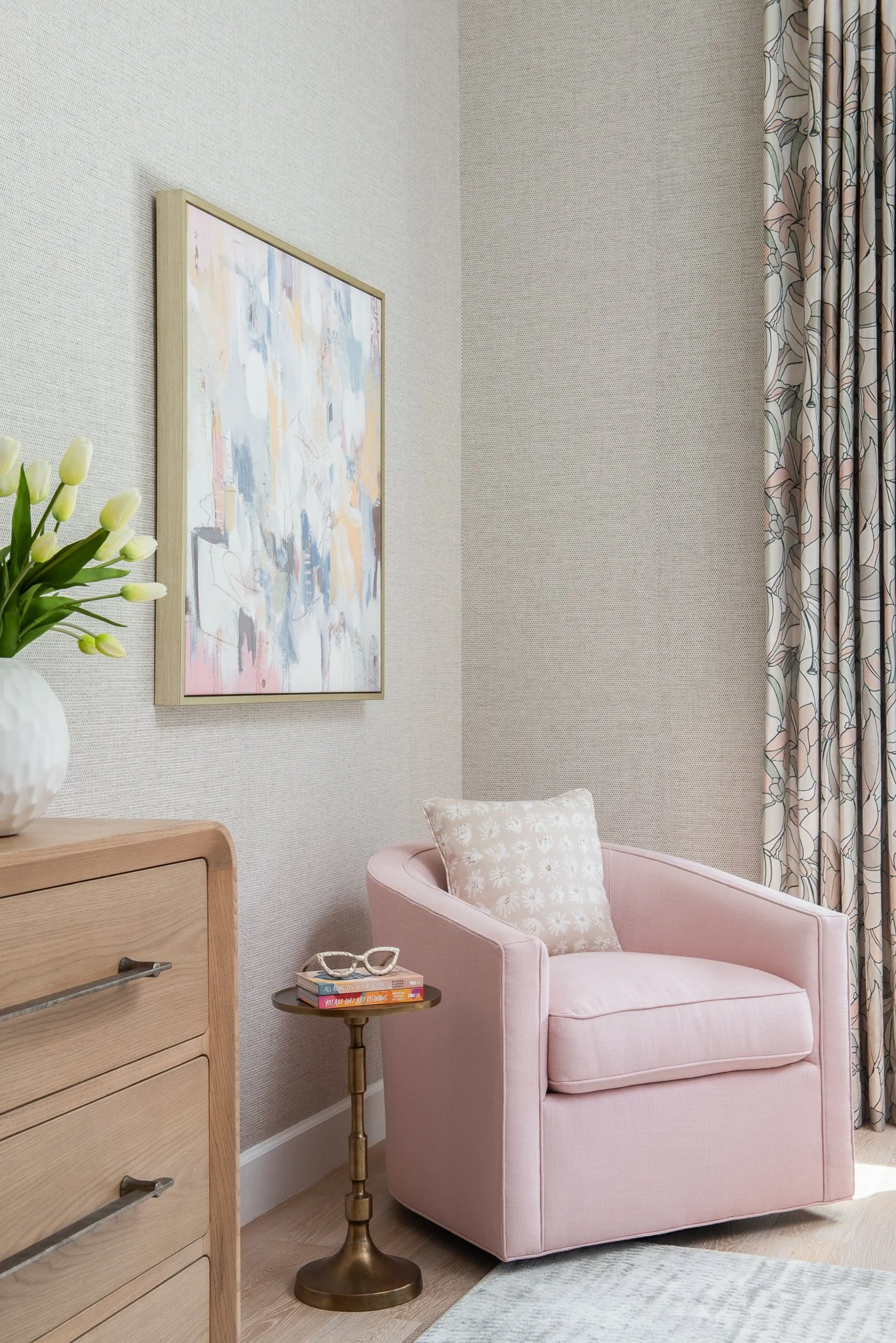 A cozy corner with a pink armchair, a cream-colored pillow with a floral pattern, a small brass side table with glasses and books, a wooden dresser with a vase of white tulips, a colorful abstract painting on the wall, long floral-patterned curtains,