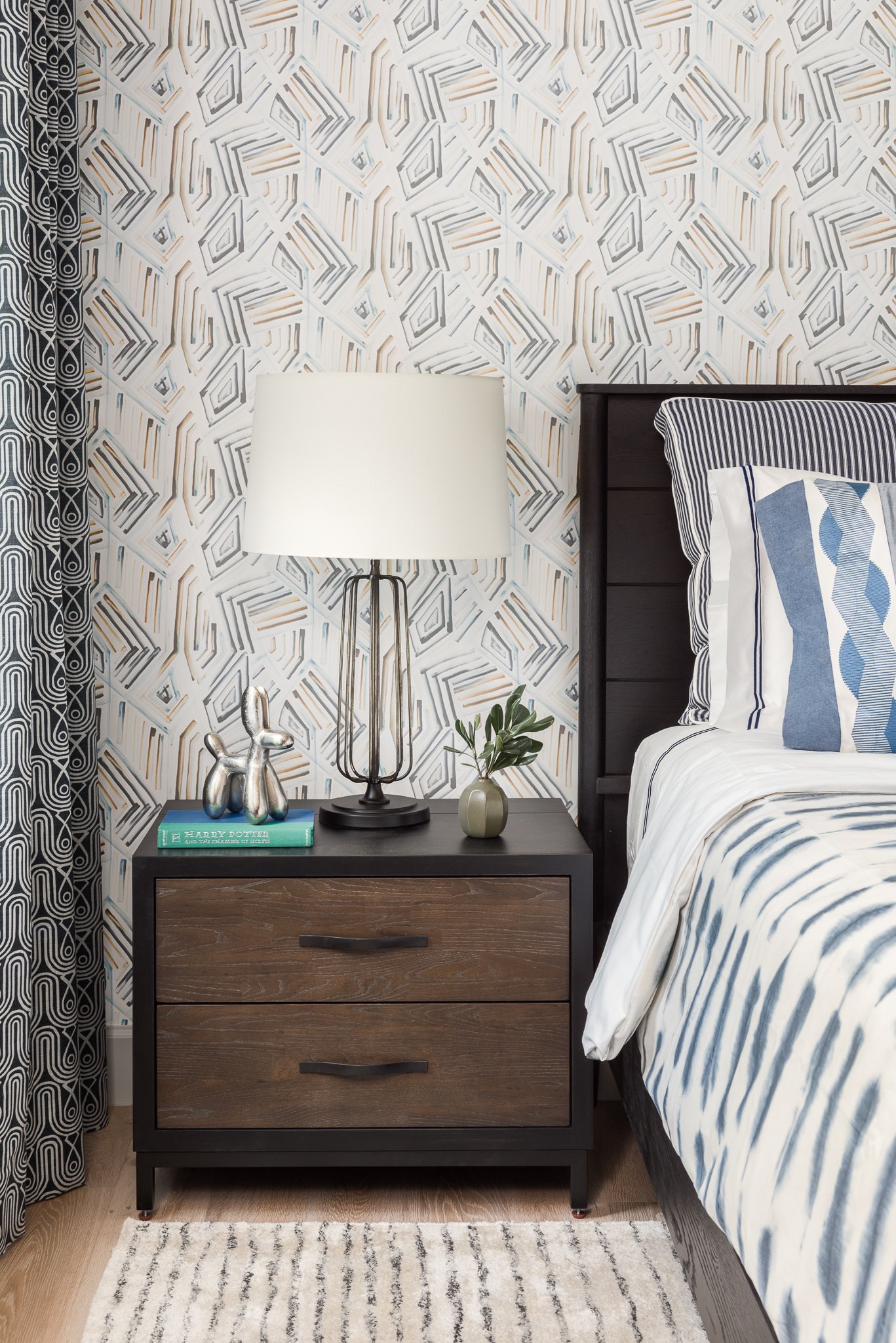 A nightstand with two drawers under a lamp with a white shade, a book titled 'Harry Potter and the College of Magic', a small silver dog figurine, and a green plant in a round vase. The background features geometric patterned wallpaper, and part of a